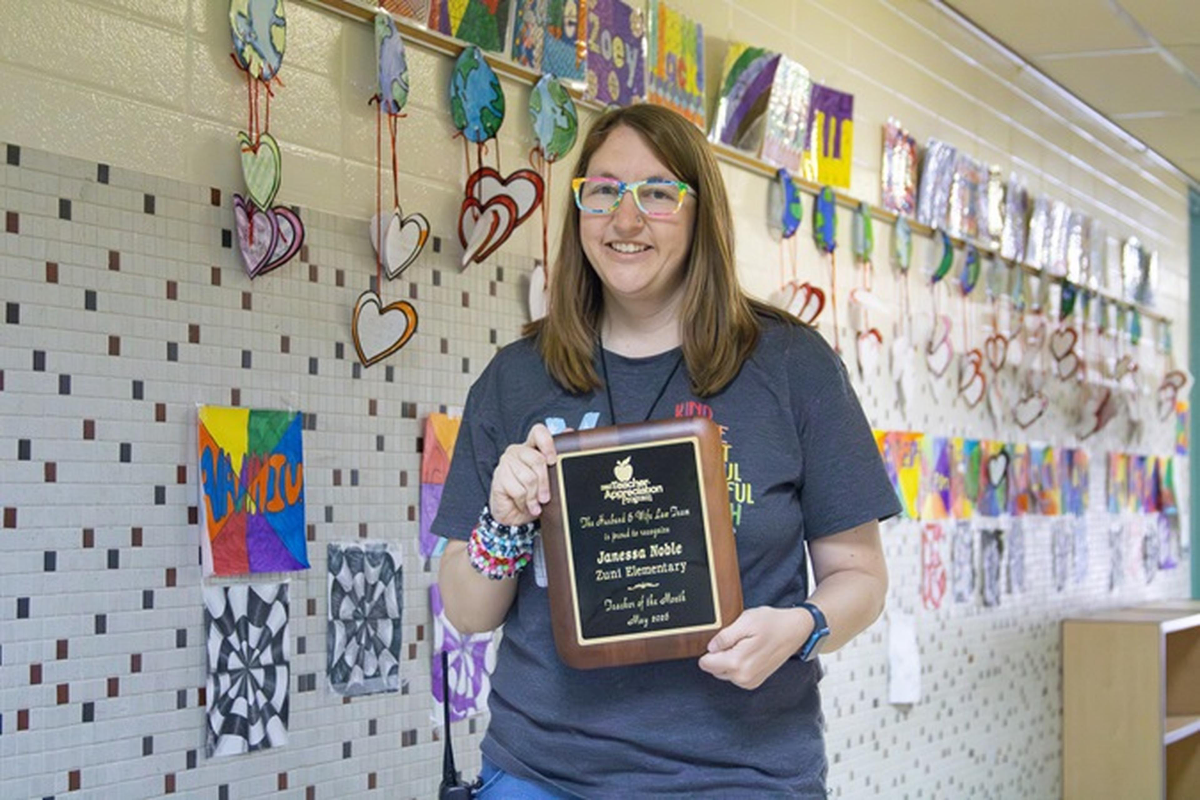 Ms. Noble holds an award plaque in a colorful school hallway decorated with hearts and student artwork.