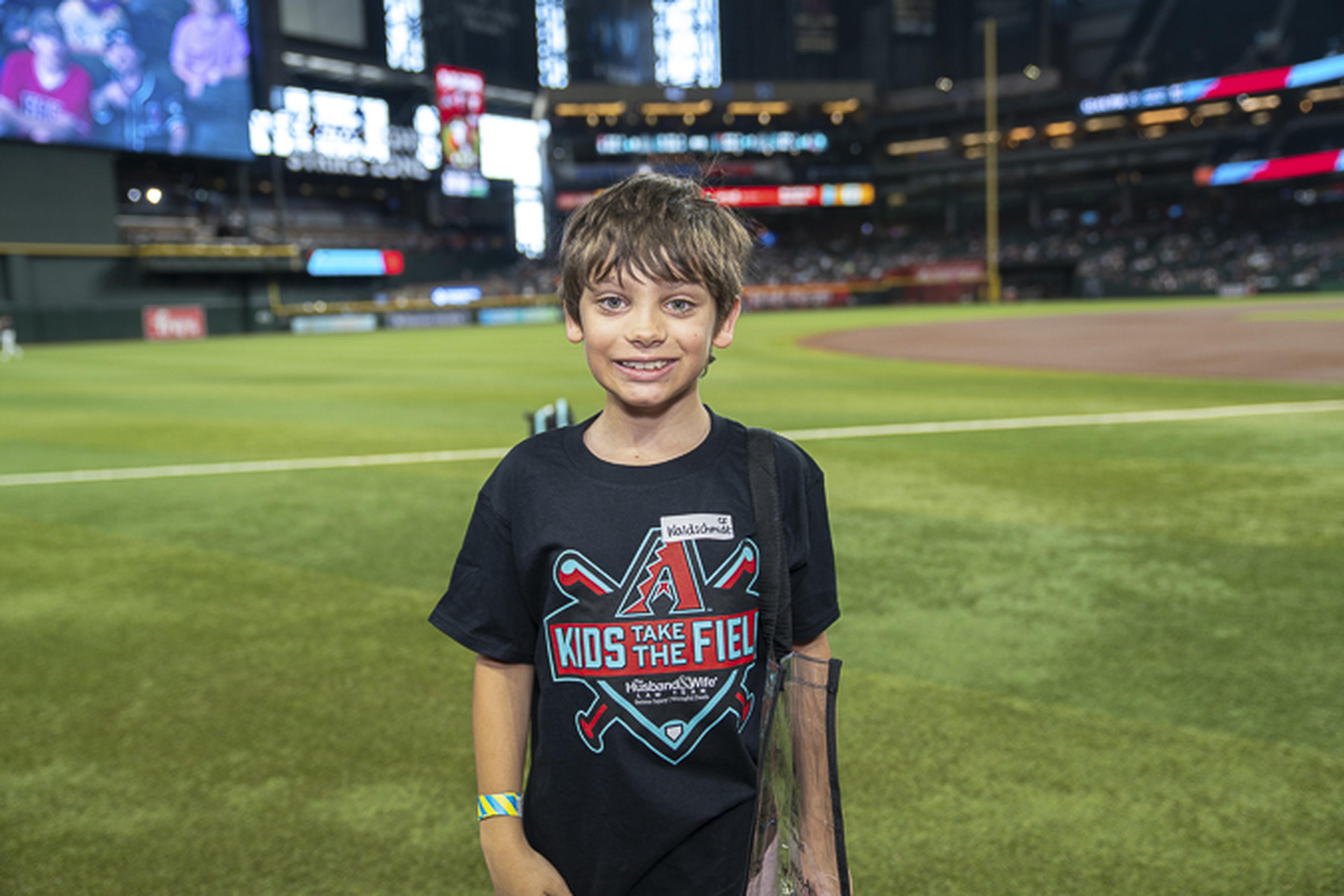 A young boy in a “Kids Take The Field” T‑shirt smiles on the baseball field with the stadium rising behind him.
