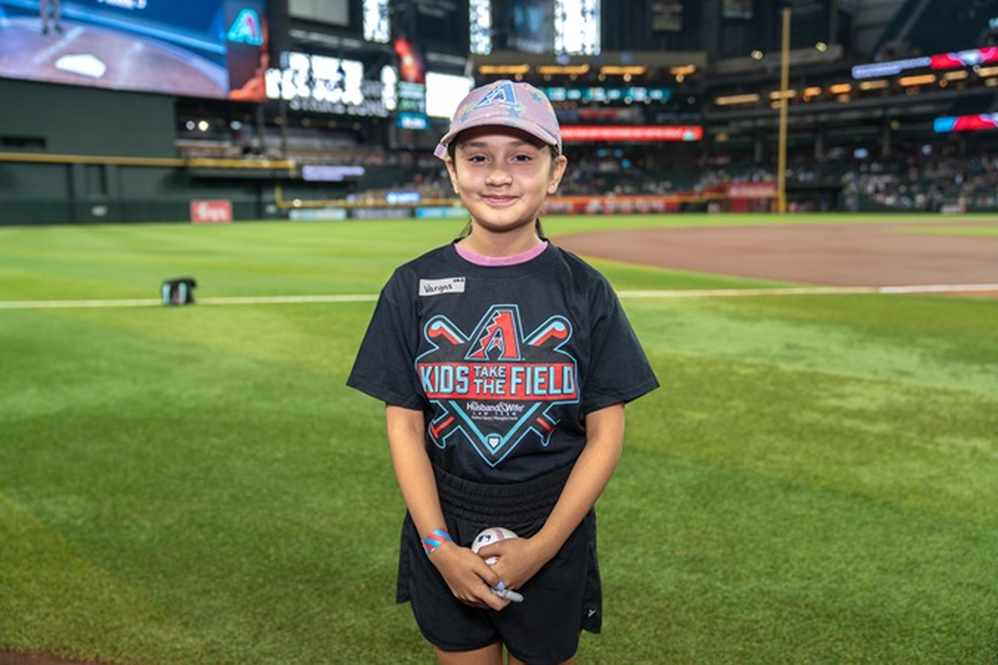 Young child in a baseball stadium, wearing a "Kids Take the Field" tee and cap, smiling on the grass near the infield.