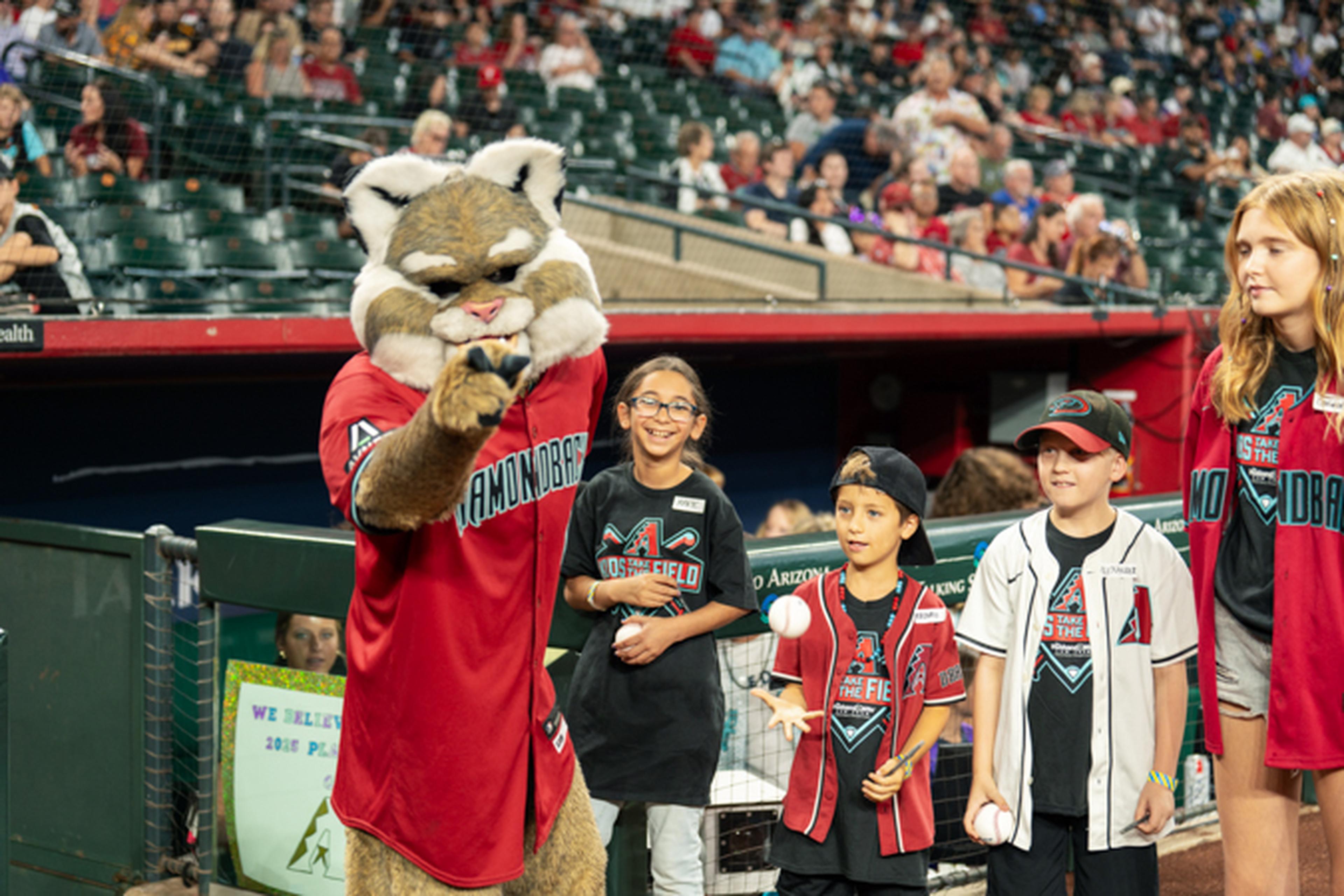 A sports mascot poses with smiling children in baseball jerseys at a stadium, with spectators in the background.