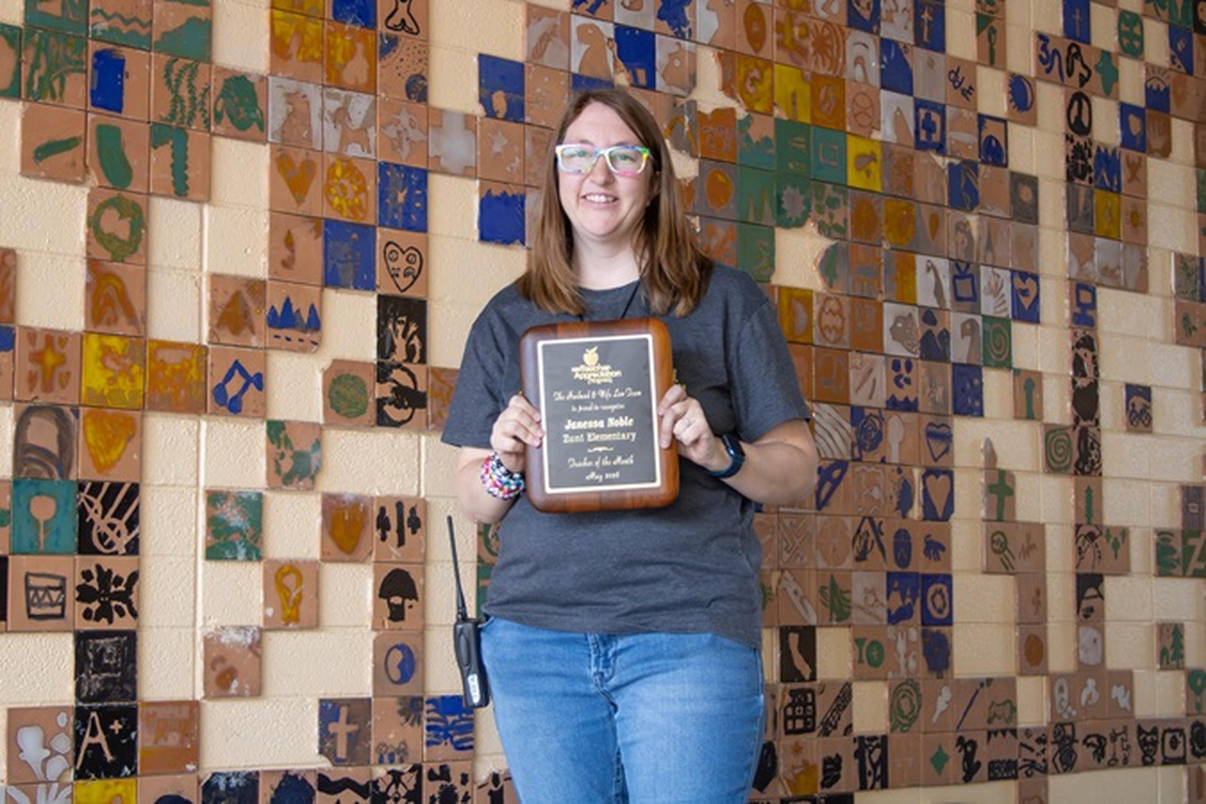 Ms. Noble smiles while holding The Husband & Wife Law Team’s Teacher Appreciation Award, standing before a colorful tiled wall.