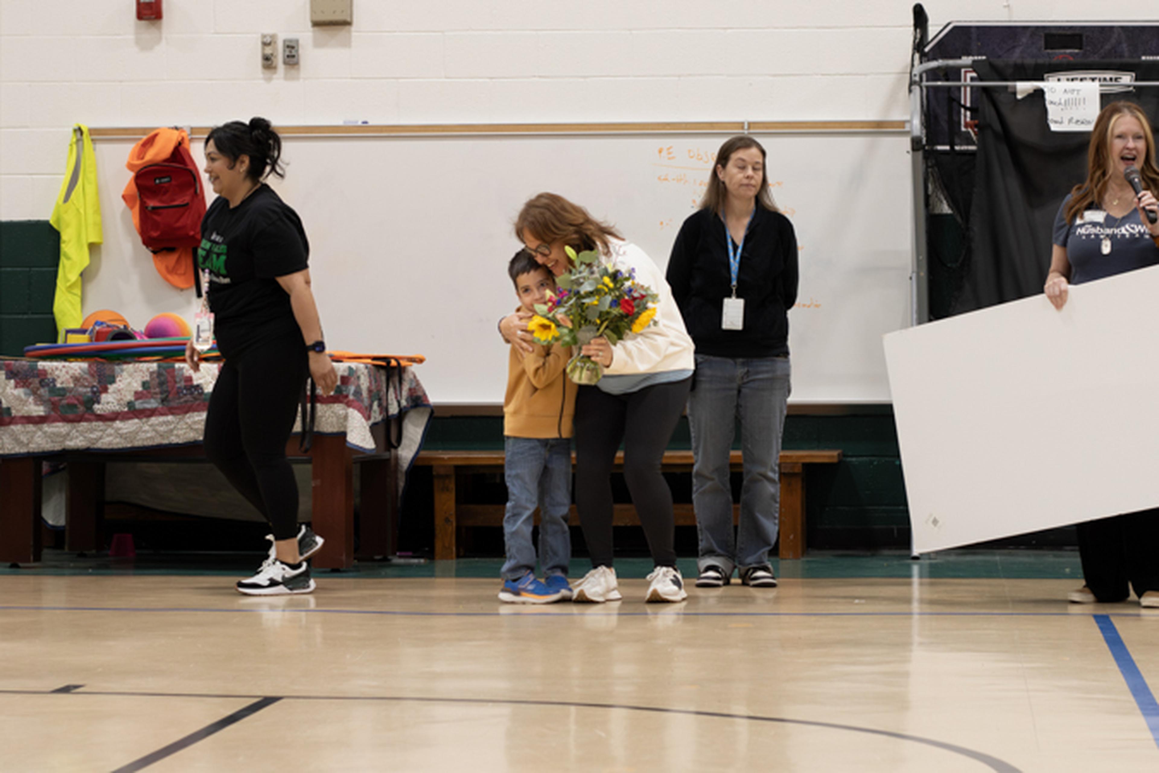 Ms. B hugs a child holding flowers in the school gymnasium, while a representative from The Husband & Wife Law Team stands nearby with a large check.
