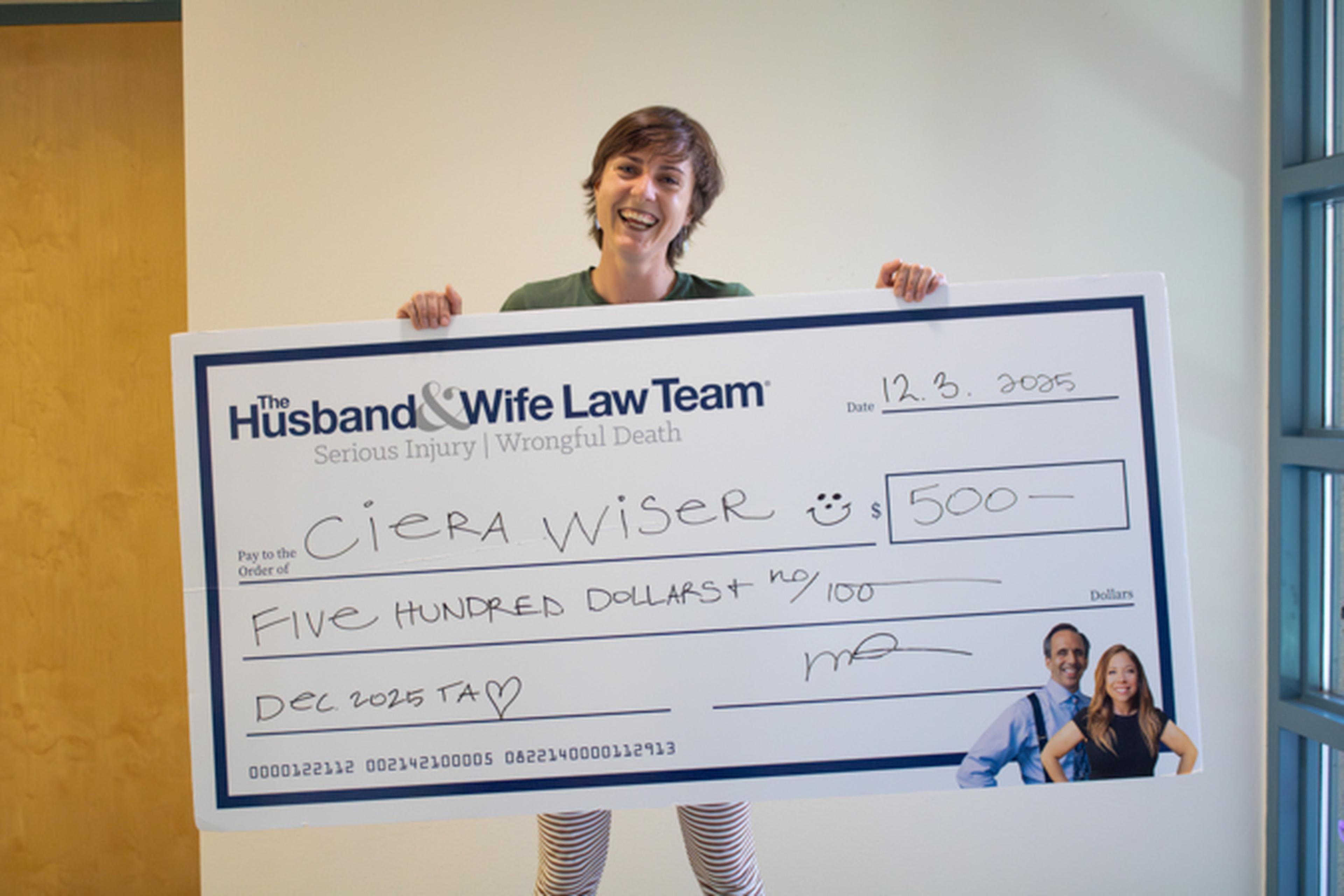 Smiling person holds oversized check from "The Husband & Wife Law Team" payable to "Ciera Wiser" for $500.