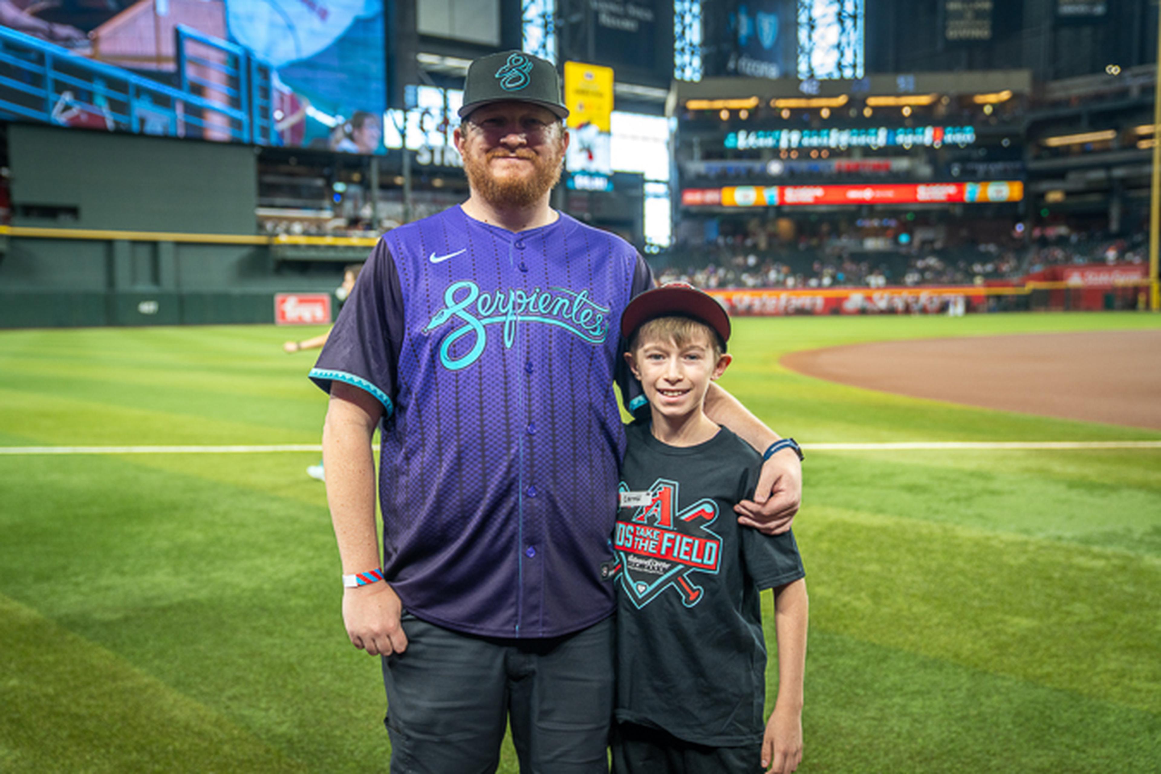 Celebrating Father’s Day, a dad in an Arizona Diamondbacks jersey and cap stands smiling on the baseball field with his son in a baseball tee.