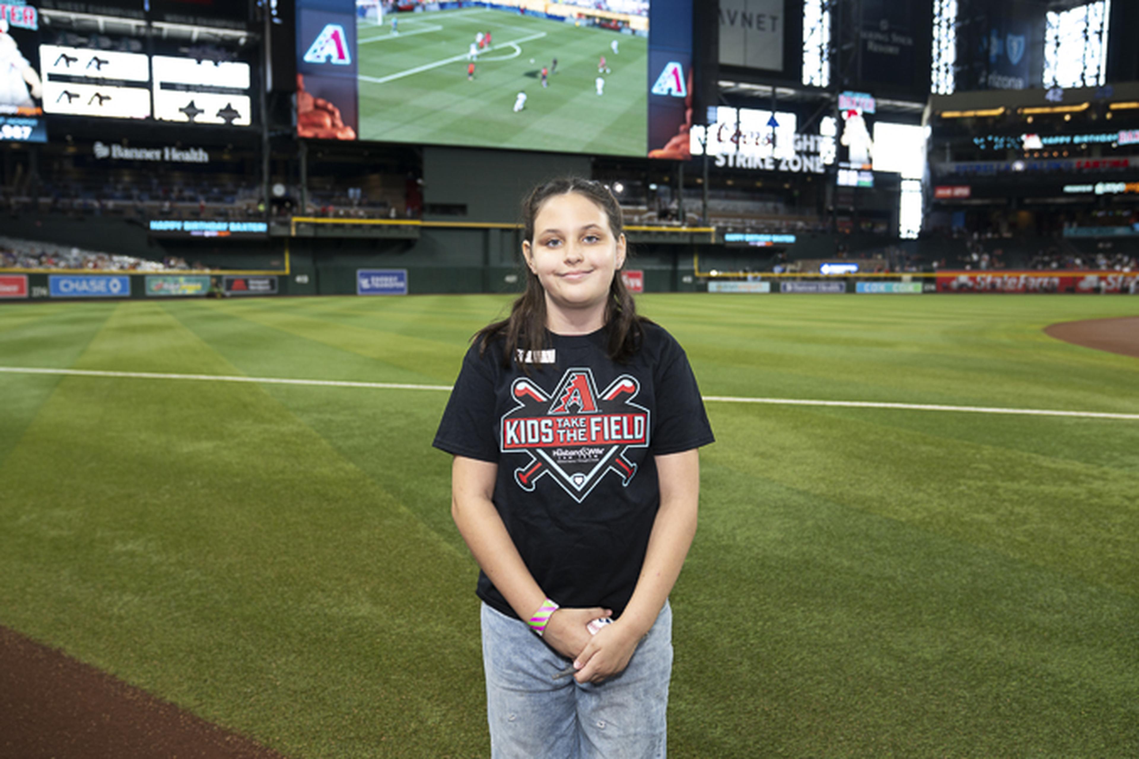 Living out a baseball dream, this young fan stands proudly on the field in her Kids Take the Field shirt, enjoying an unforgettable experience made possible by The Husband & Wife Law Team