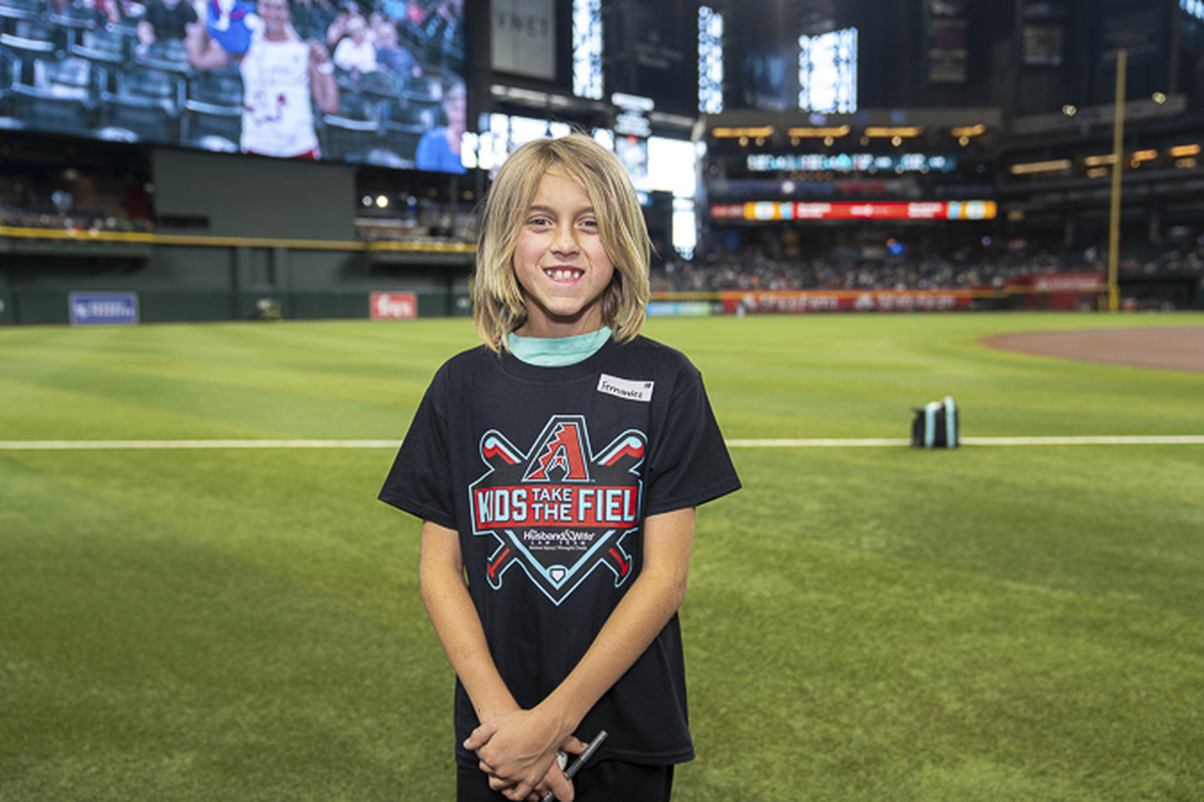 A happy young fan wearing a “Kids Take the Field” tee poses on the field, the stadium and cheering crowd in the background.