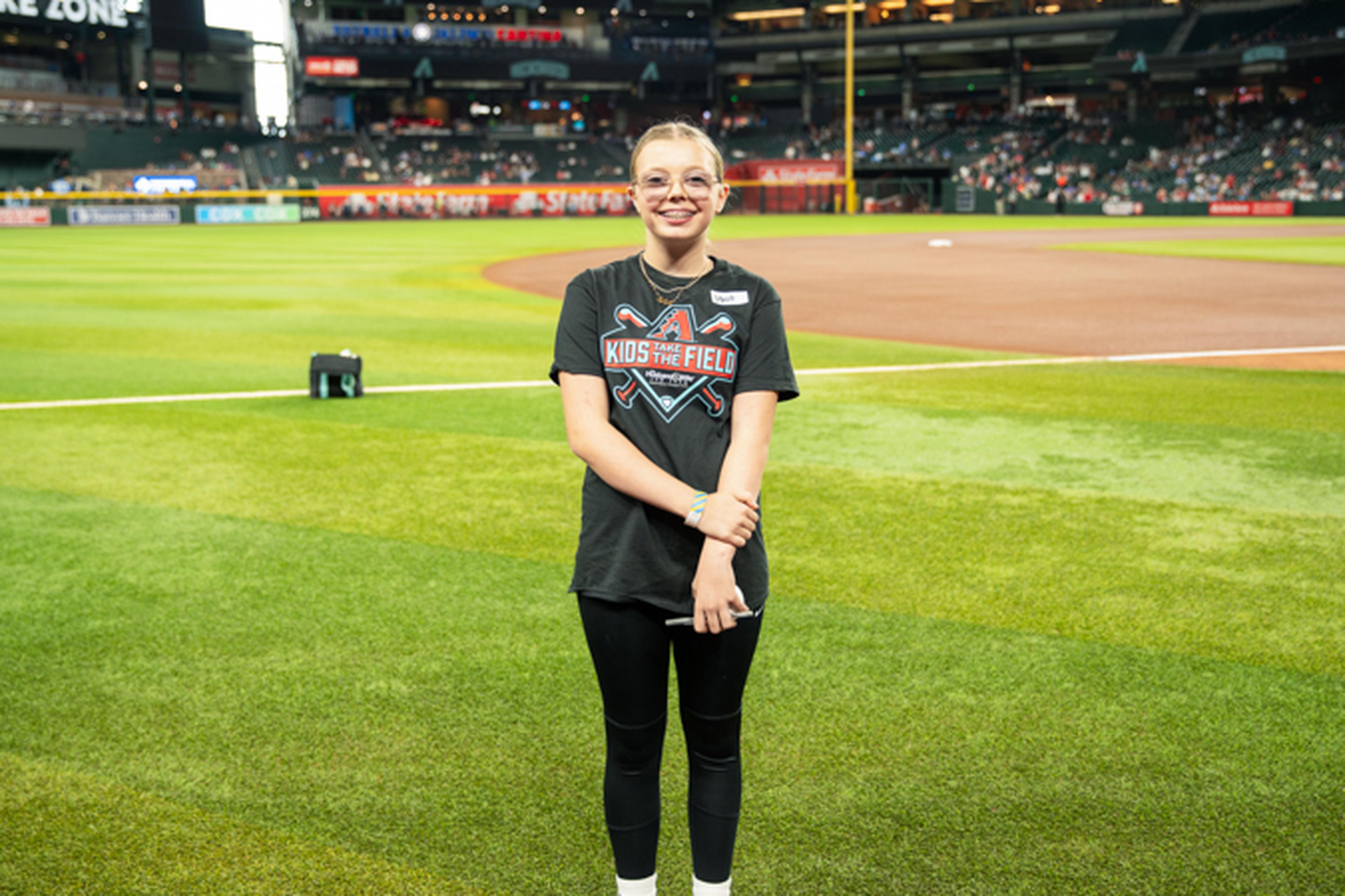 A person stands on a baseball field wearing a "Kids on the Field" shirt, smiling with arms crossed. Stadium seats and field visible in the background.
