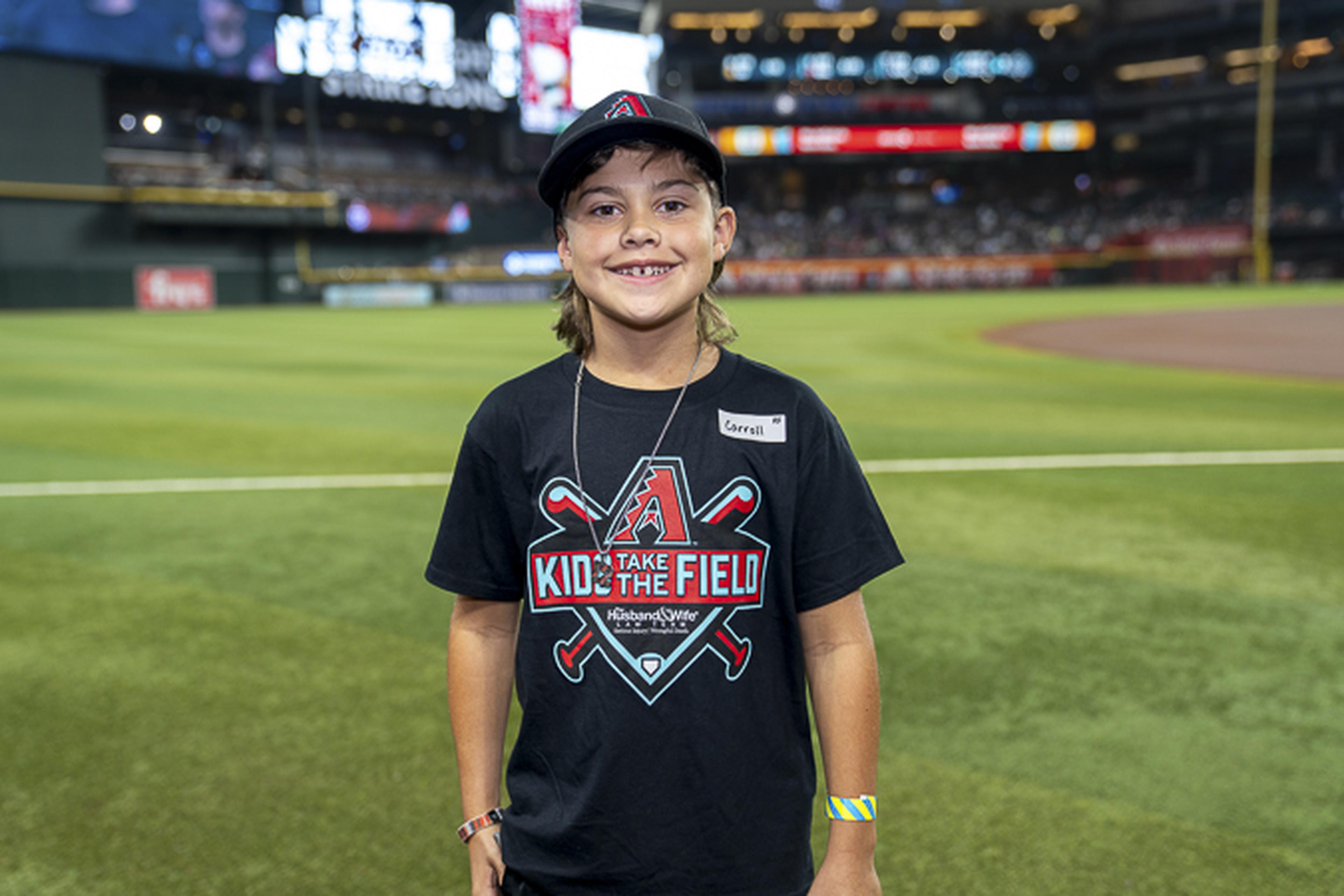 Smiling beneath the bright stadium lights, a young fan in a “Kids Take the Field” tee and cap stands on the field with the crowd in the background.