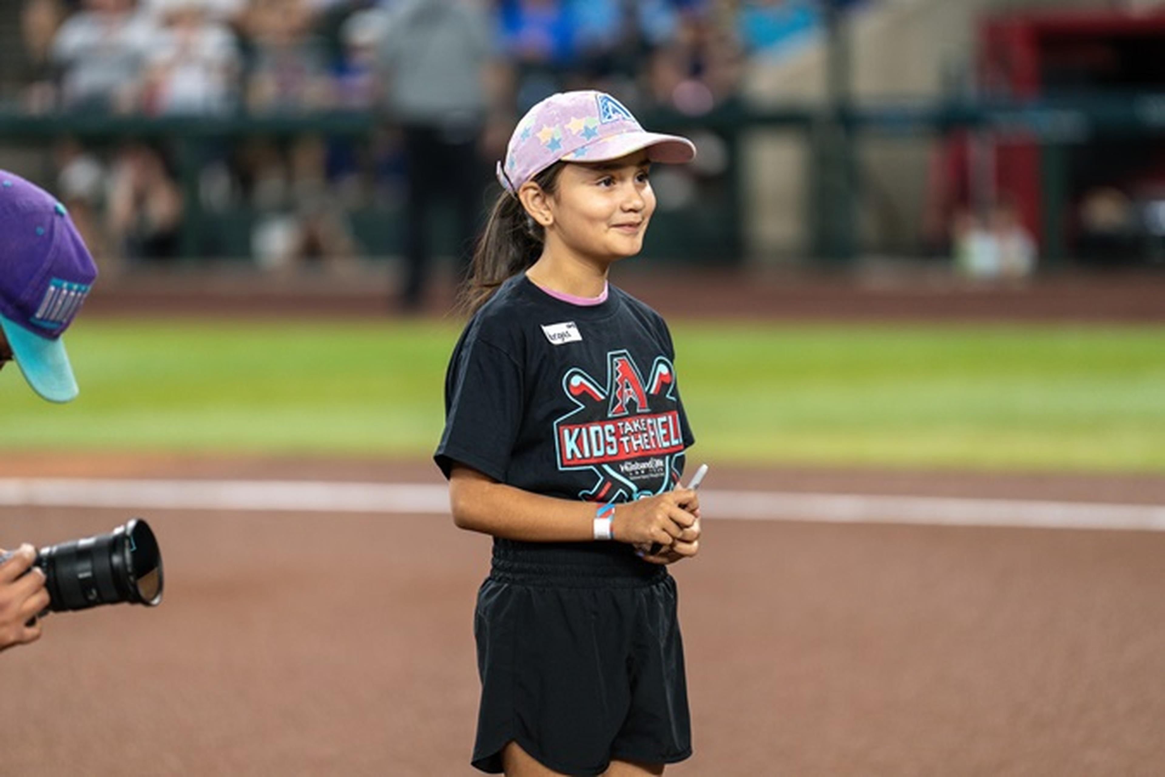 Girl in a pink cap and black shorts stands on a baseball field, holding a pen, with a crowd in the background.