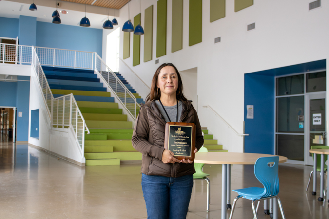 Ms. Kim, our Santa Fe Teacher of the Month! Woman standing in a bright school lobby holding an award plaque, with colorful tiered seating, stairs and blue chairs in the background.