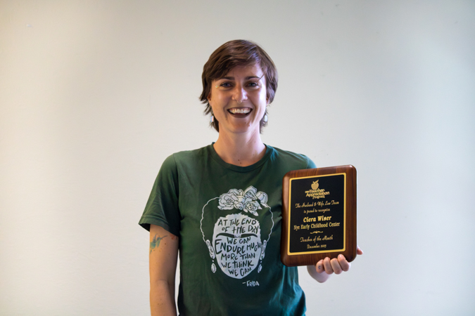 Ms. Ciera smiles with her Teacher of the Month plaque Smiling short-haired woman in green graphic T-shirt holding a wooden award plaque against a plain light background.