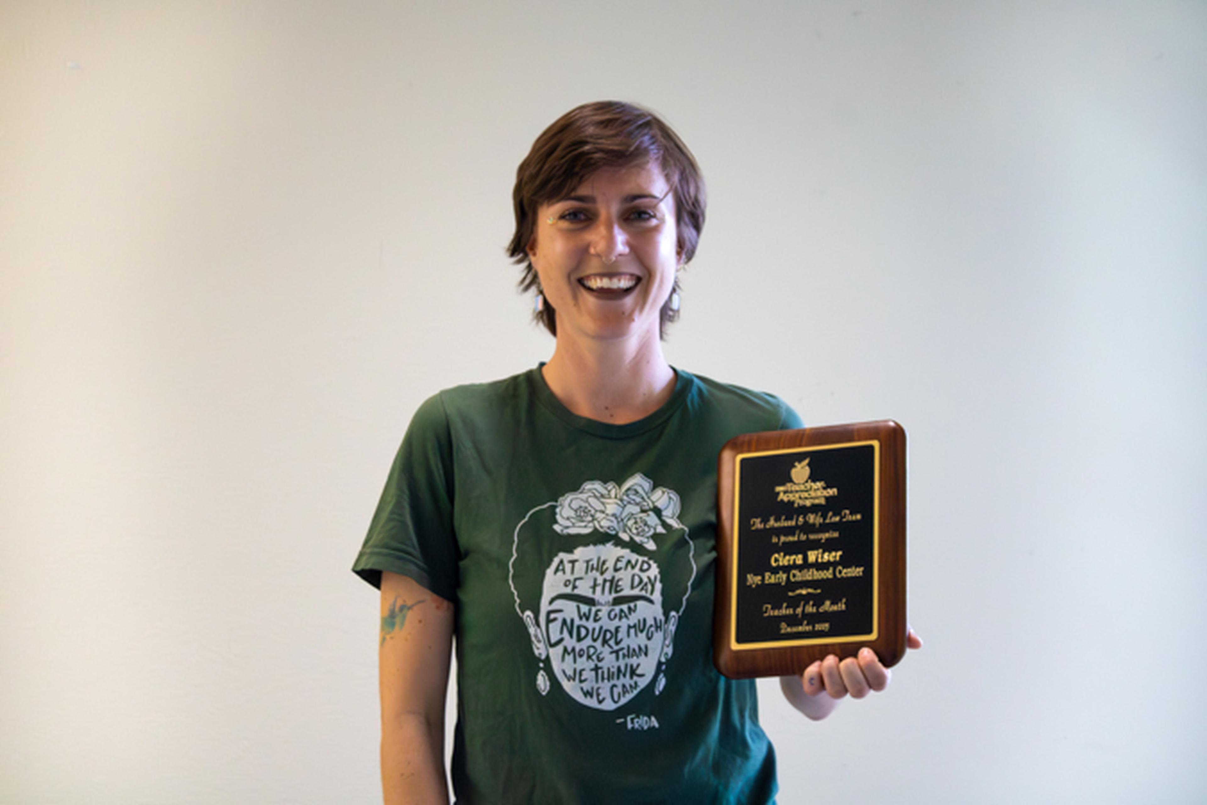 Smiling short-haired woman in green graphic T-shirt holding a wooden award plaque against a plain light background.