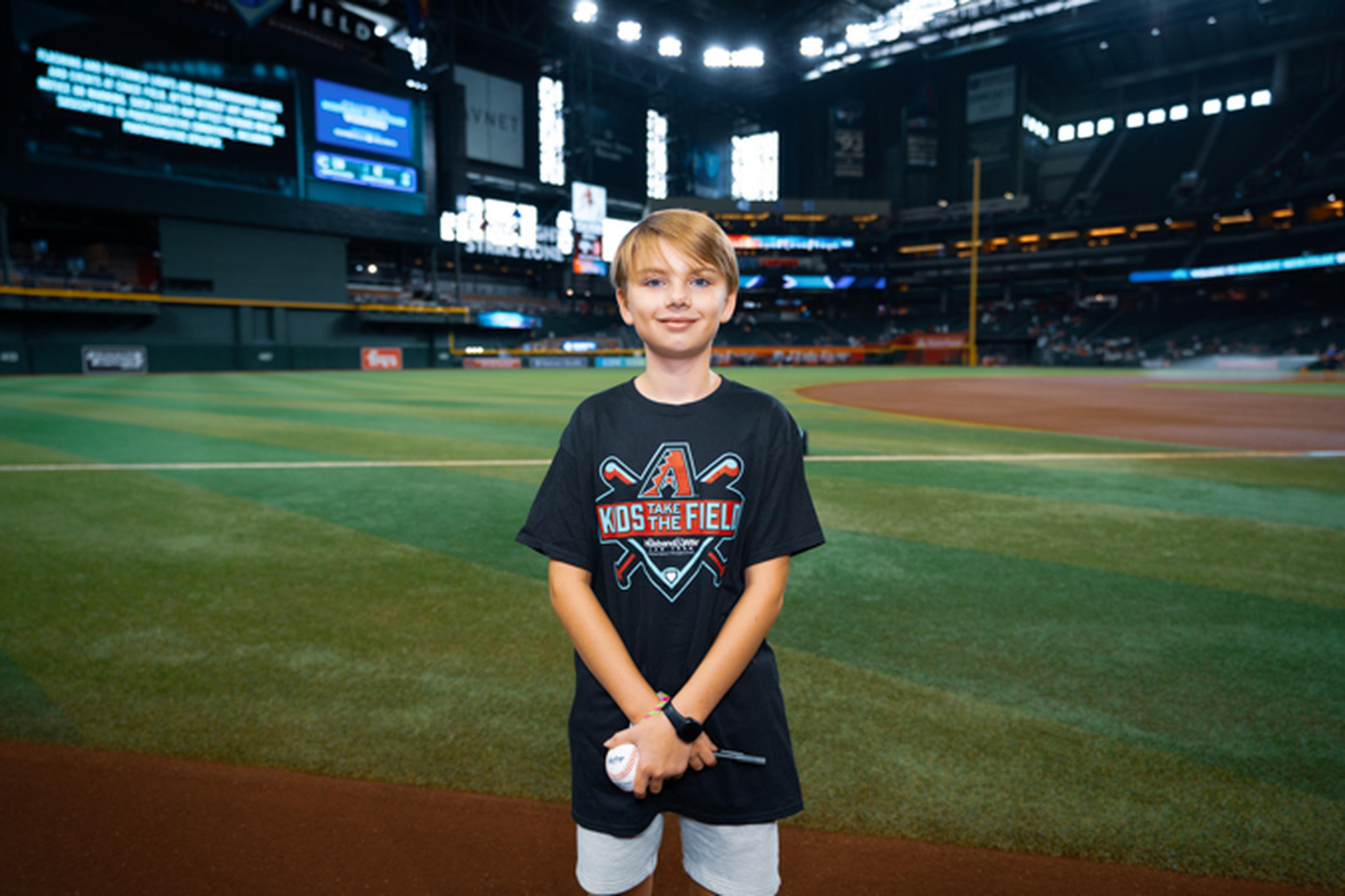 Young person standing on a baseball field, holding a ball, wearing a "Kids on the Field" shirt, with an empty stadium in the background.