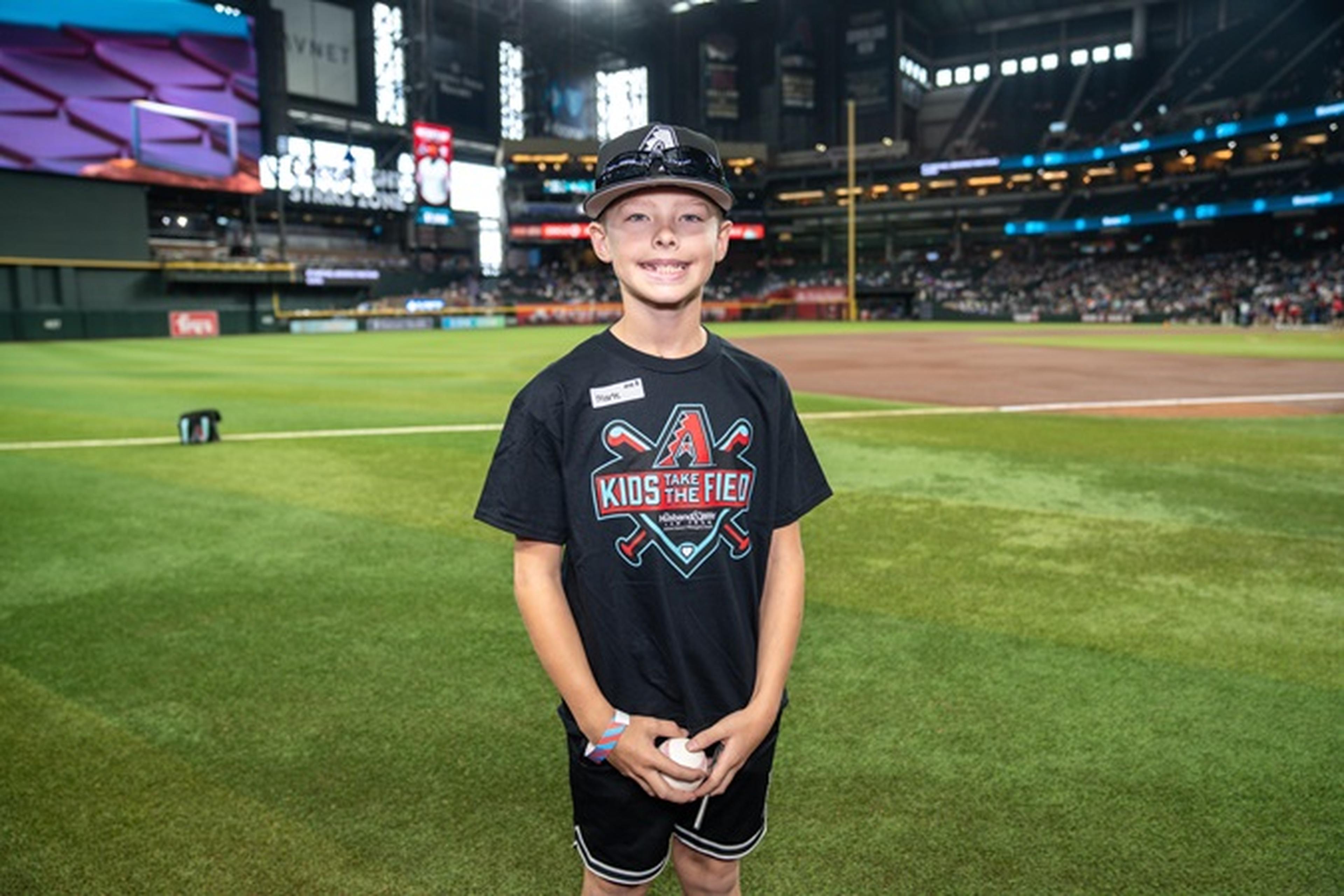 Young boy wearing a "Kids Take the Field" T-shirt and cap, smiling and holding a baseball on a stadium field with fans in the background.