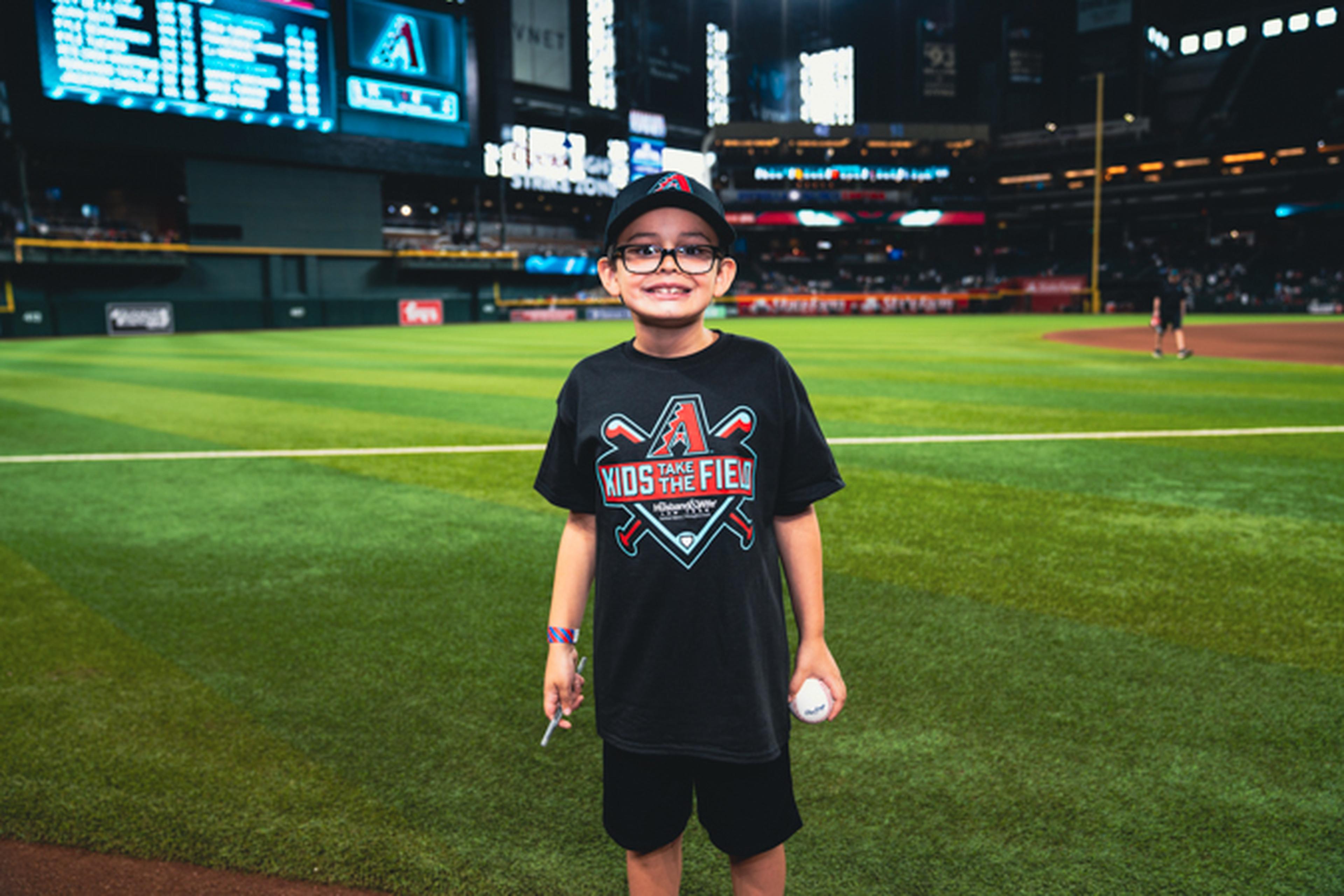 A smiling child in a baseball cap and "Kids on the Field" shirt stands on a baseball field holding a ball, with a stadium in the background.
