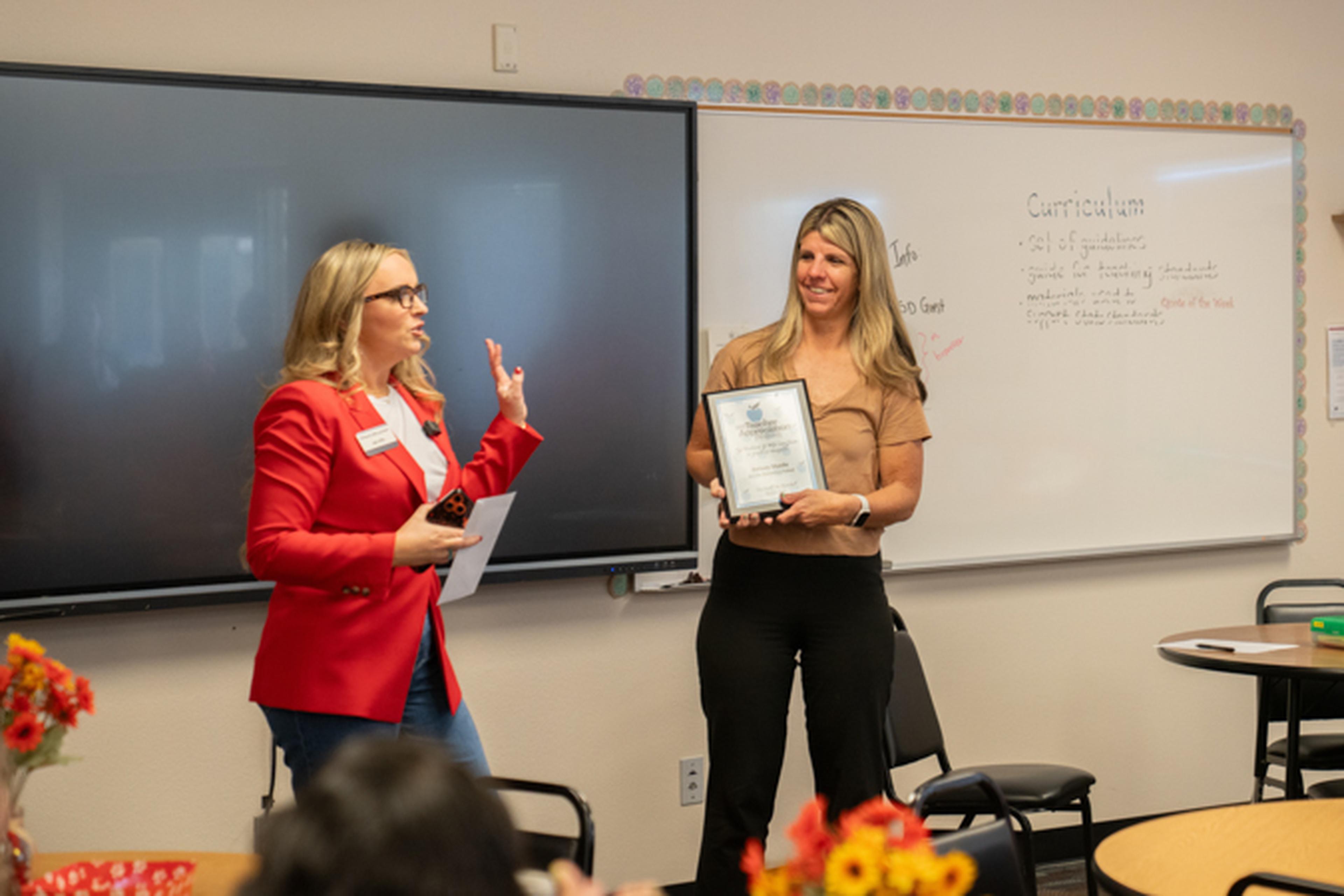 Jen from The Husband & Wife Law Team presents a framed certificate to Ms. Shade, who smiles. Flowers are on a table in the foreground.