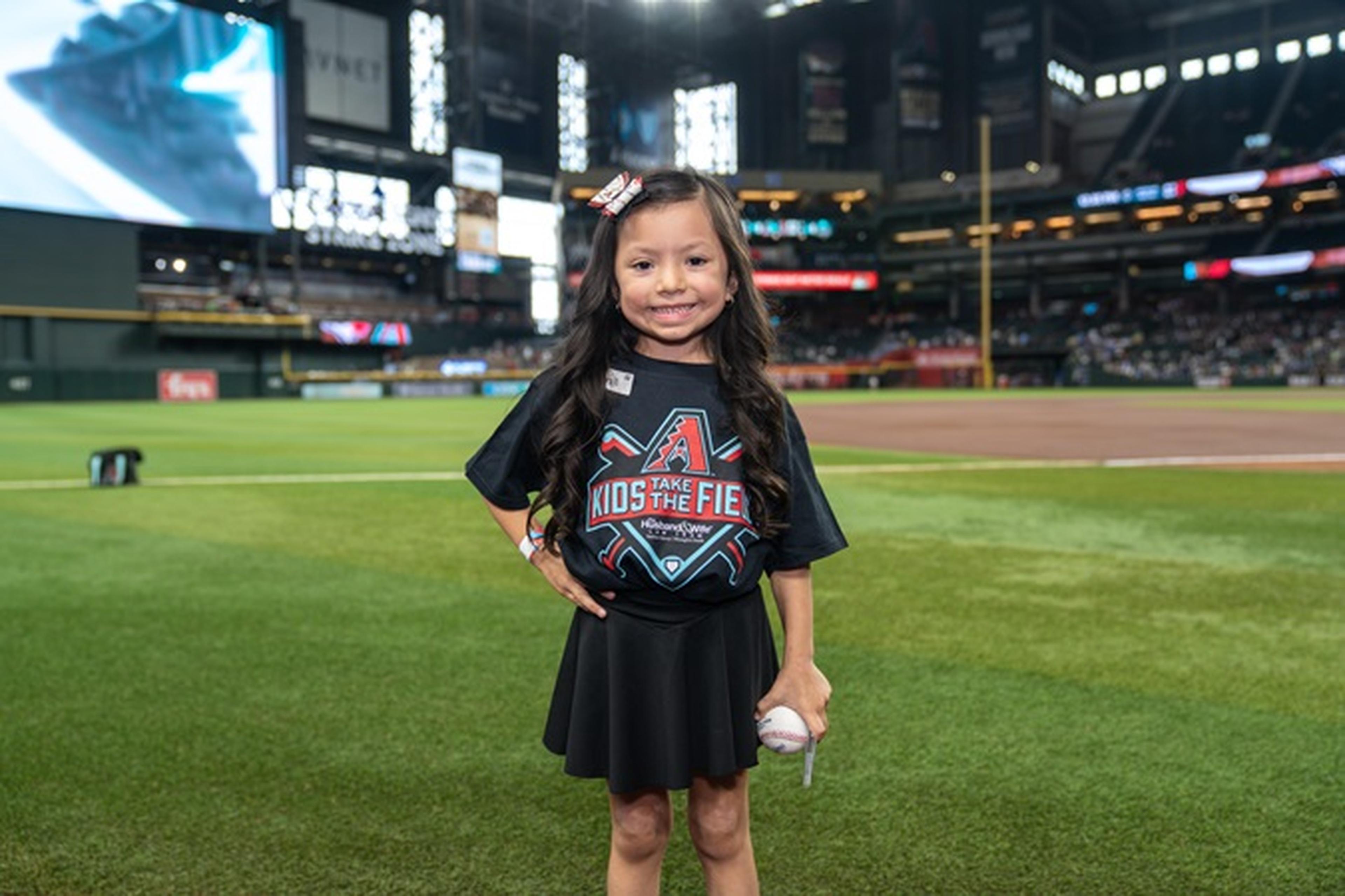 Young girl standing on a baseball field, smiling, holding a ball, wearing a "Take Kids to The Field" T-shirt. Stadium lights and empty stands behind.