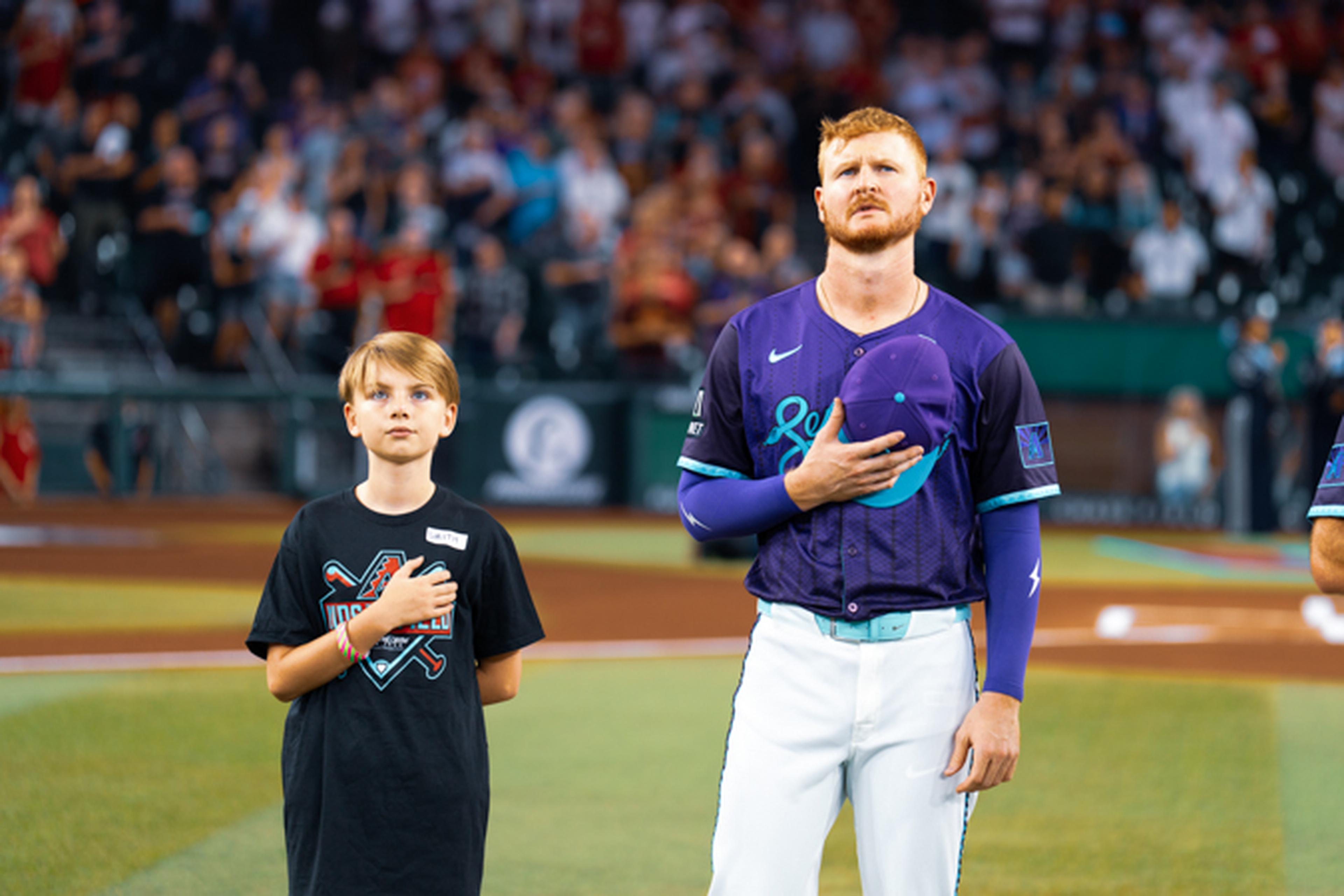 A baseball player and a young boy stand with hands over hearts on a field, surrounded by a blurred crowd.