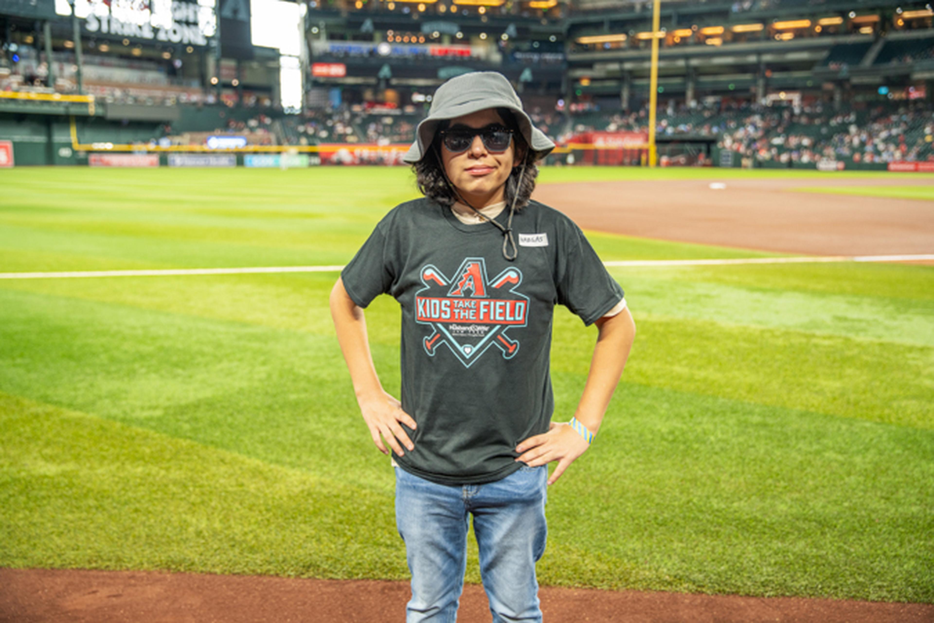 A person in a "Kids Take the Field" shirt and sun hat stands on a baseball field, with hands on hips and a stadium in the background.