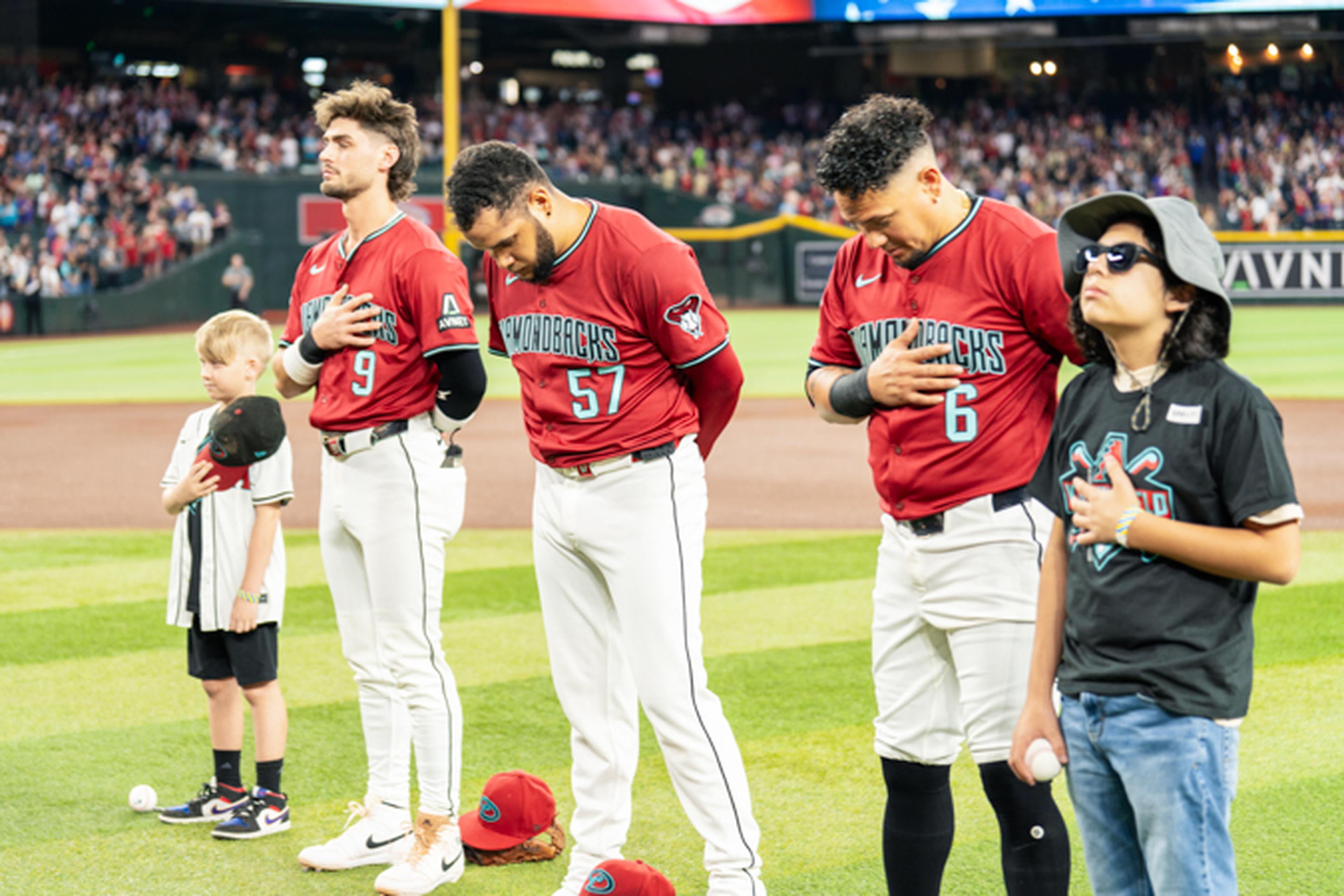 Baseball players and two children stand on a field with hands over hearts during a national anthem ceremony.