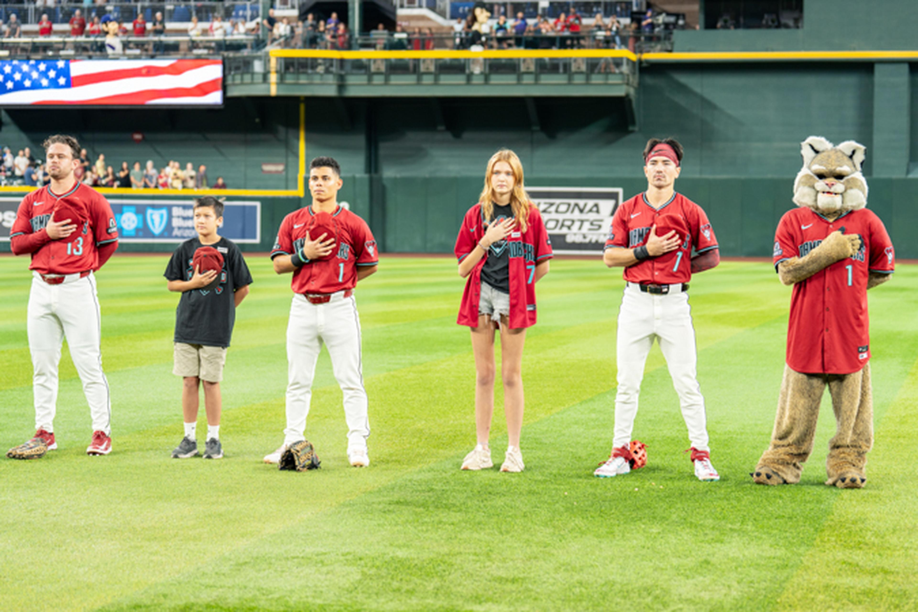 Baseball players, a young boy, a girl, and a mascot stand with hands on hearts on a field, with an American flag in the background.