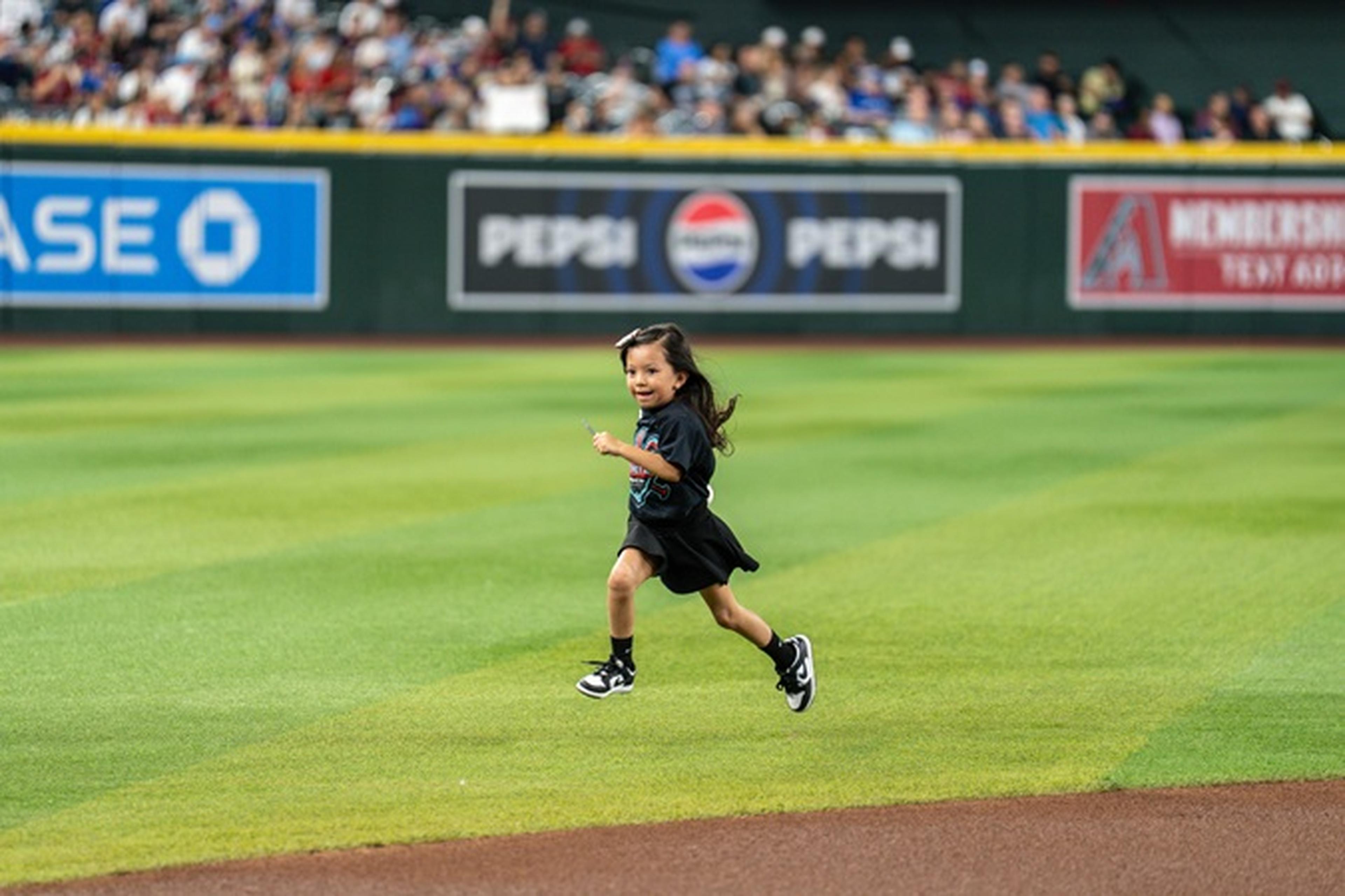 Young child joyfully running on a baseball field, with spectators and advertising banners in the background.