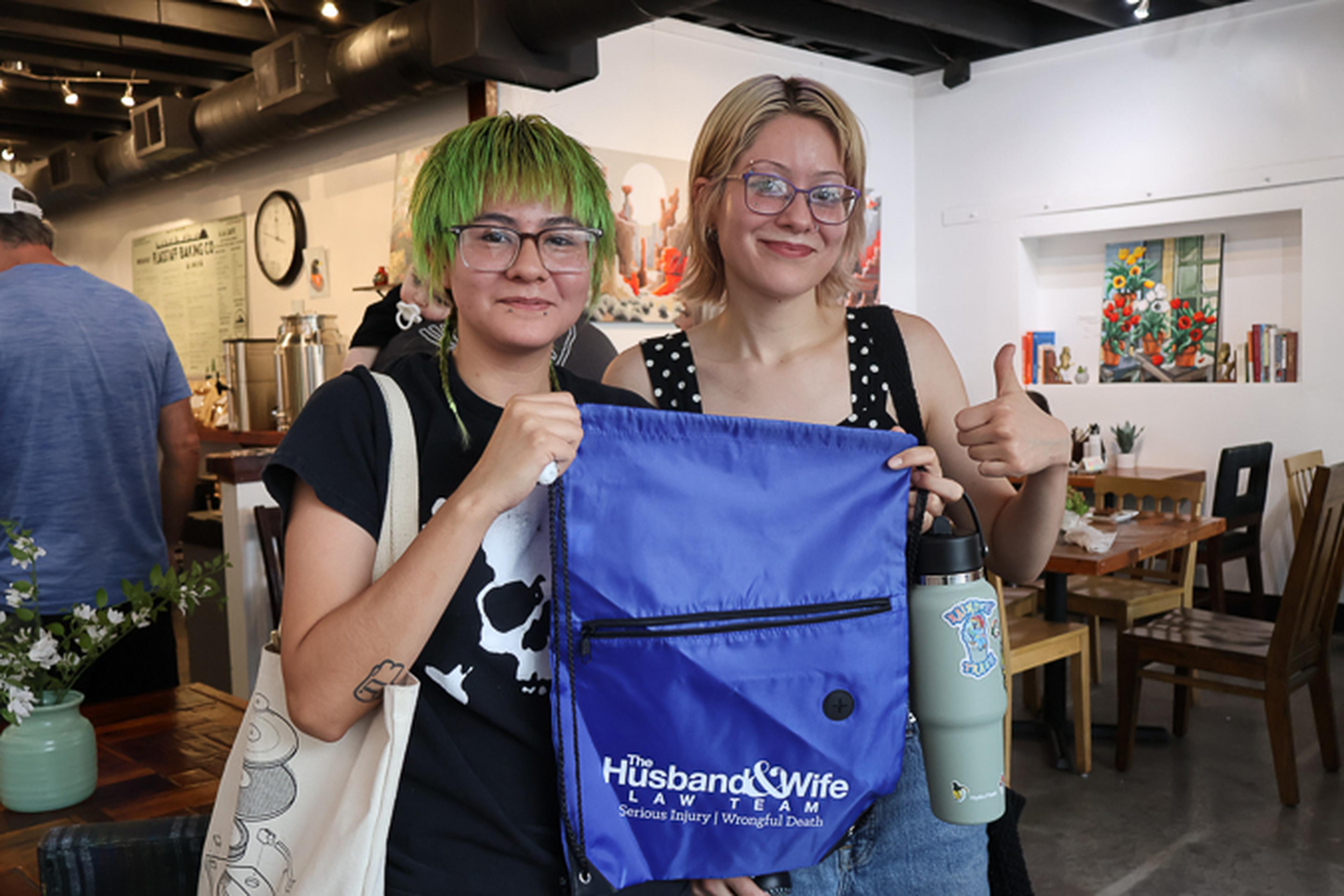 Two attendees show off their Husband & Wife Law Team drawstring bag while enjoying a community event at Flagstaff Baking Co., complete with smiles and a thumbs-up.
