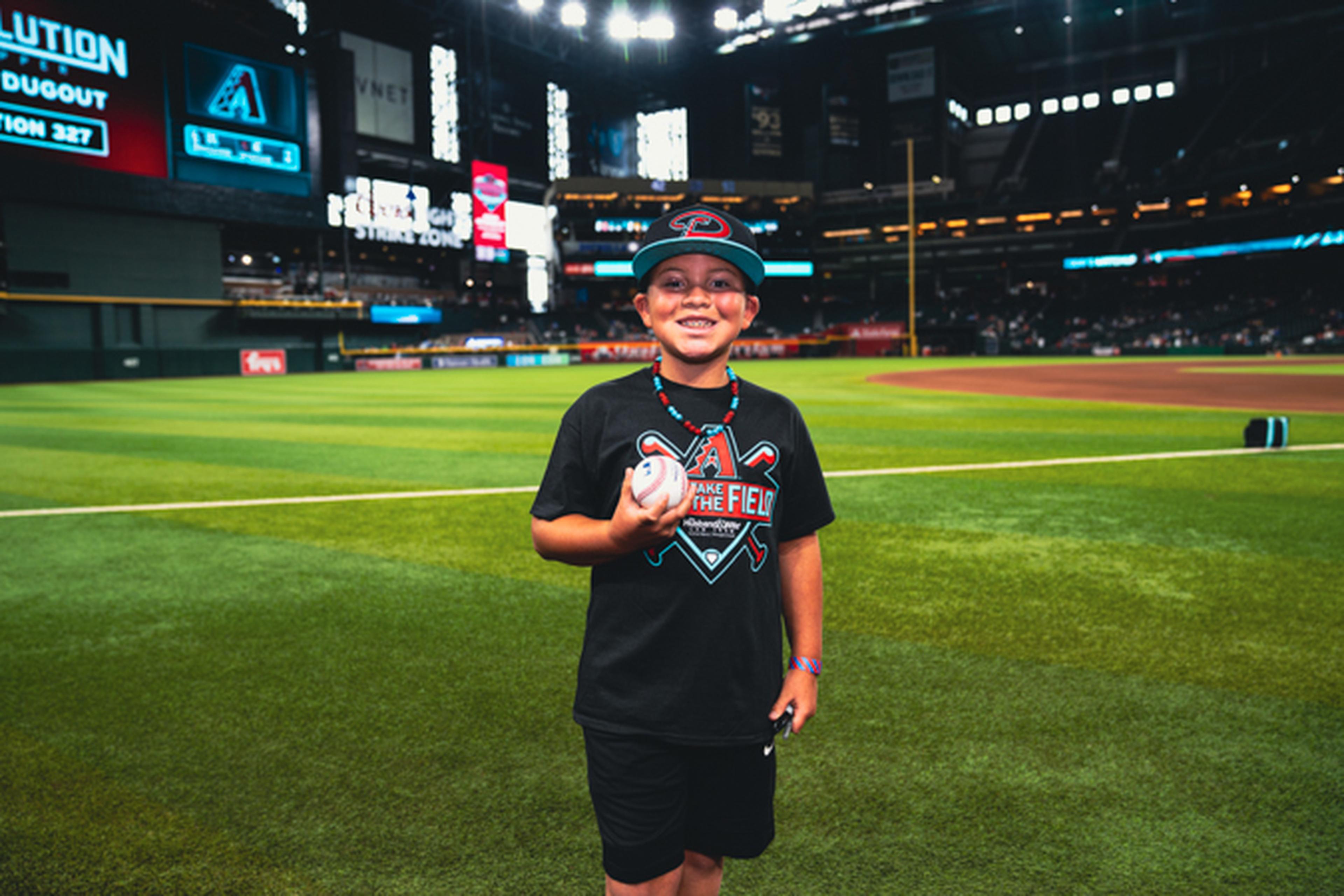 Young boy in baseball cap and glasses holds a baseball, standing on a professional field with stadium lights and screens in the background.