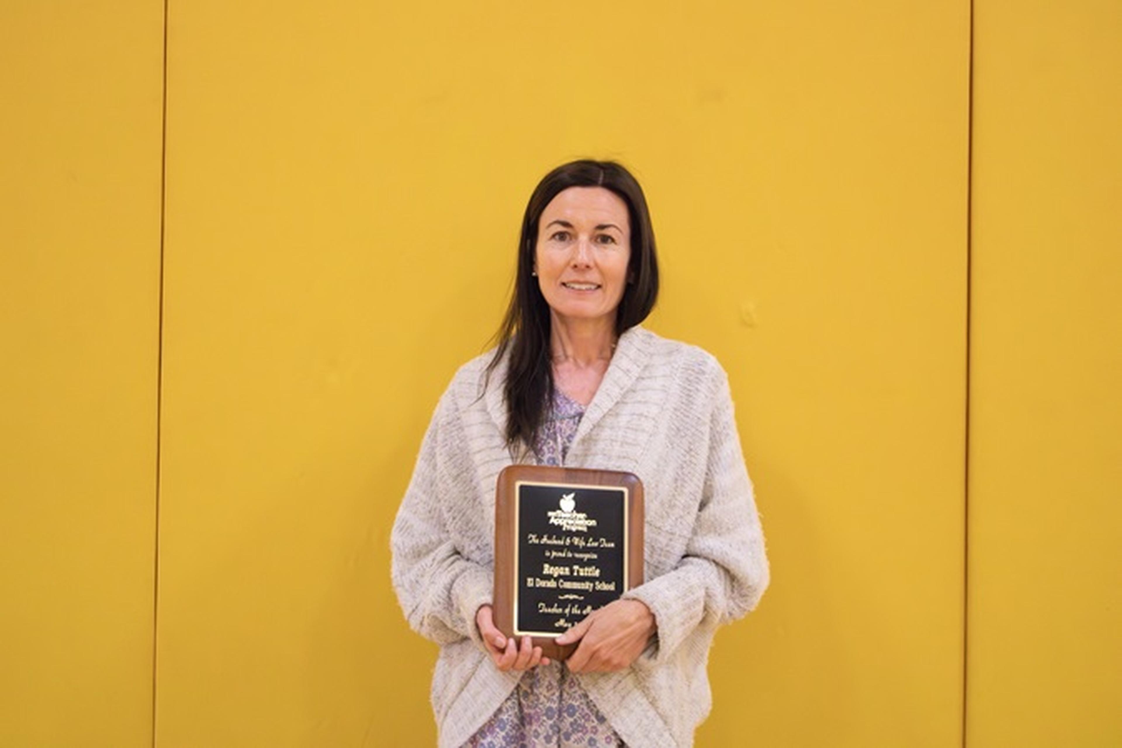 Ms. Tuttle smiles at the camera while holding The Husband & Wife Law Team’s Teacher Appreciation Award, standing against a yellow backdrop in a light cardigan and floral dress.