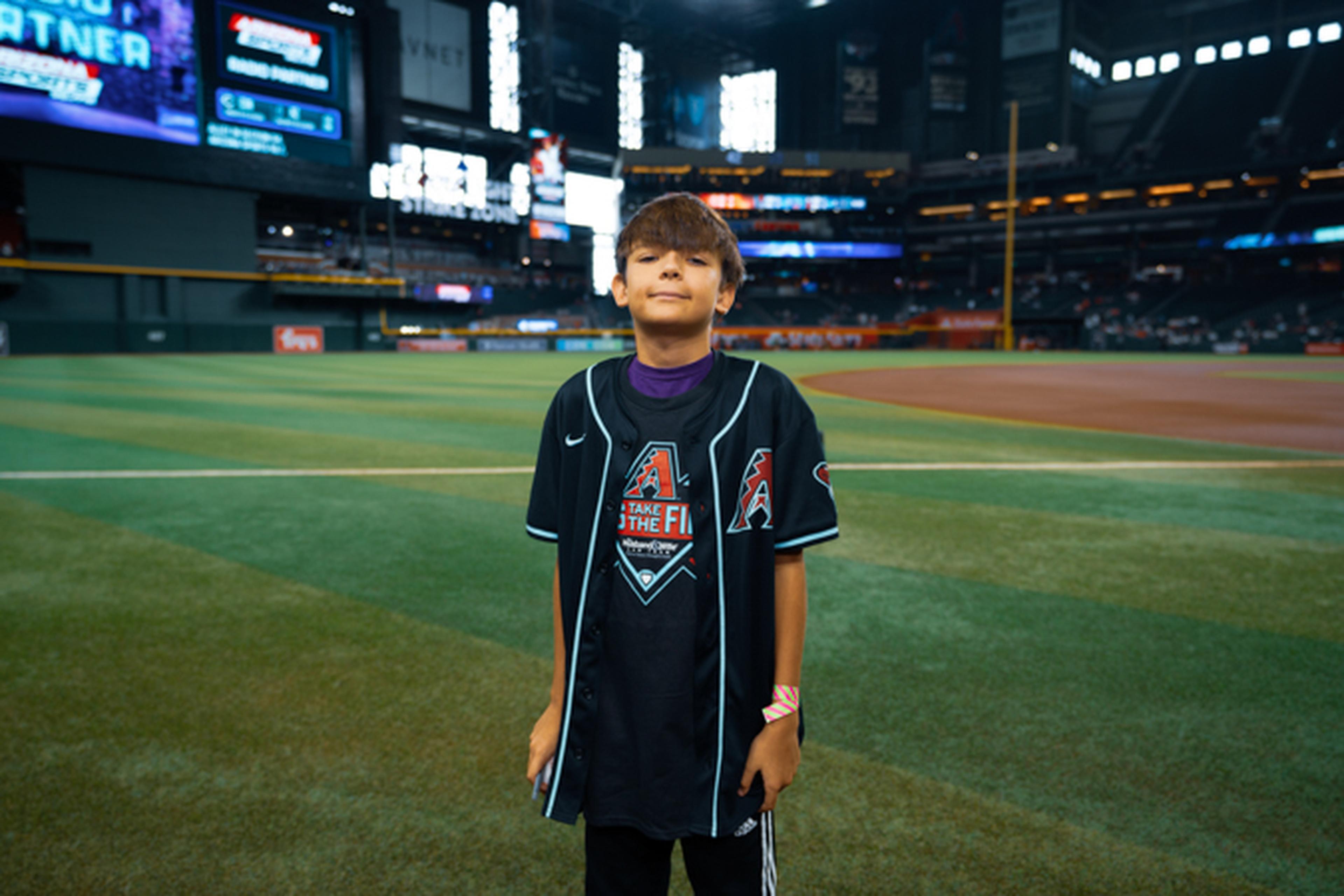 A young person stands on a baseball field wearing a black jersey with a logo, surrounded by an empty stadium.