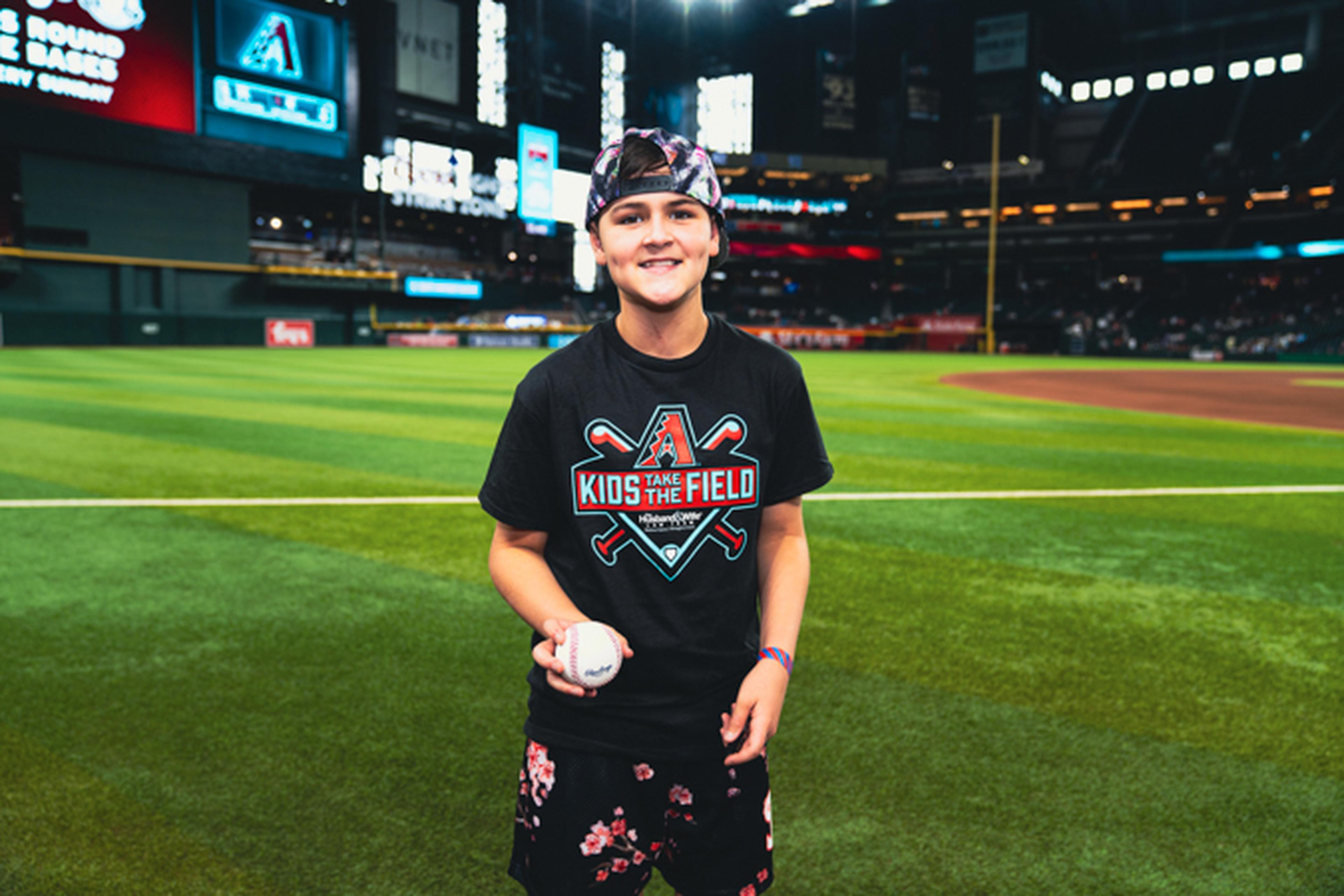 Young person on a baseball field, wearing a cap and a "Kids on the Field" shirt, holding a baseball, with a stadium backdrop.