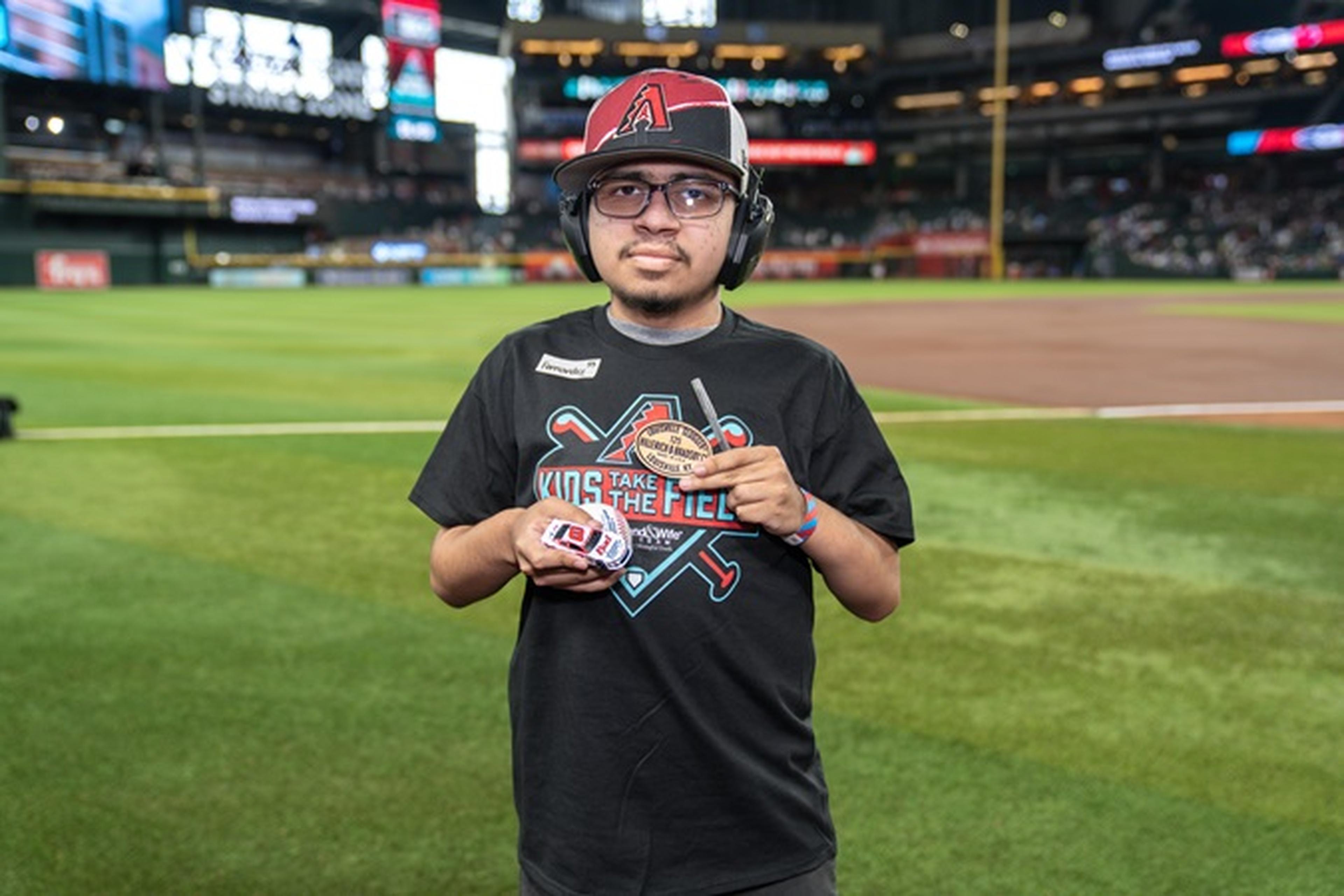 A person wearing a baseball cap and headphones stands on a grassy baseball field, holding items while facing the camera.
