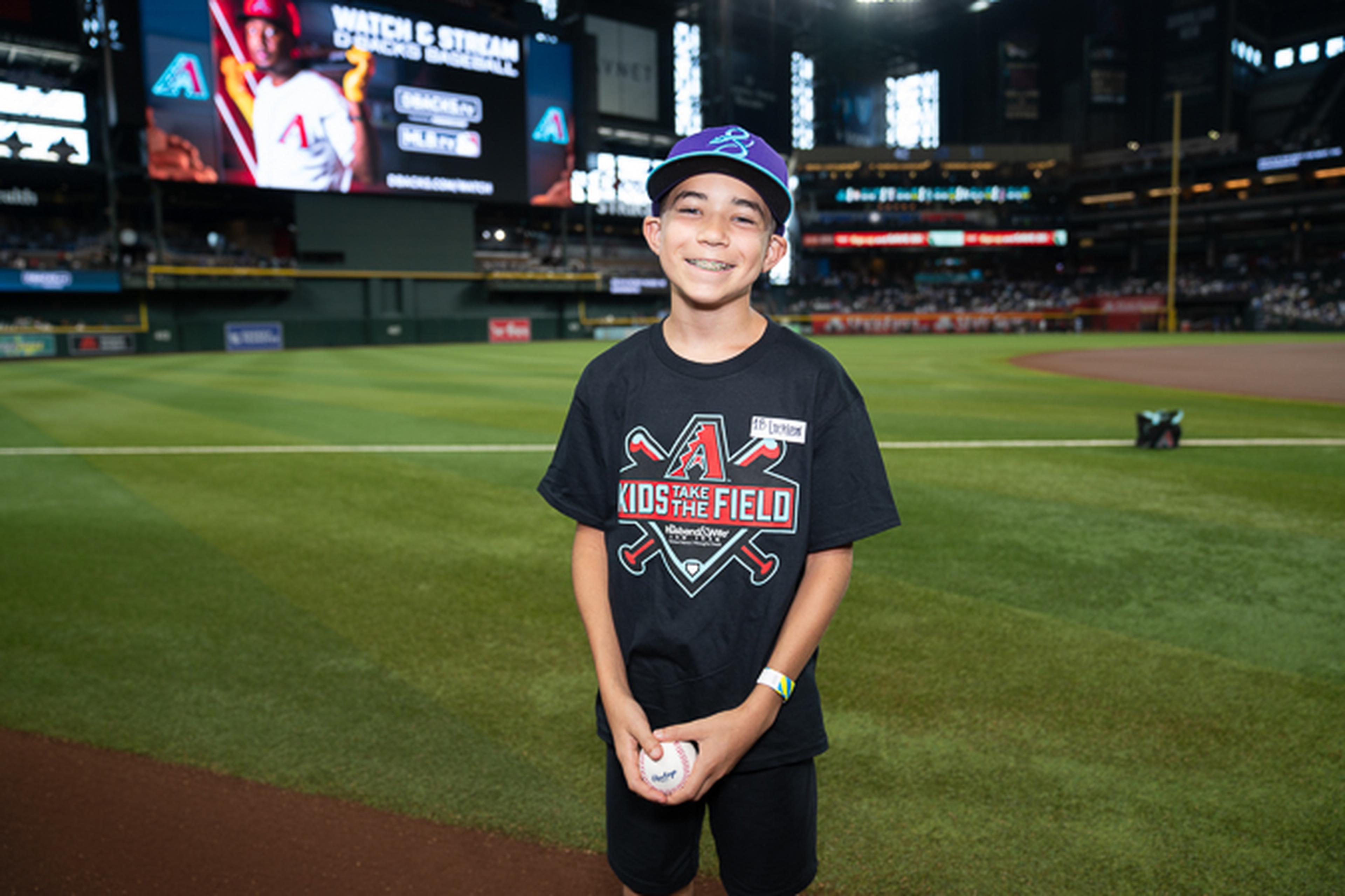 A young Arizona Diamondbacks fan proudly holds a baseball on the field at Chase Field, enjoying a memorable day at the ballpark surrounded by fellow fans.