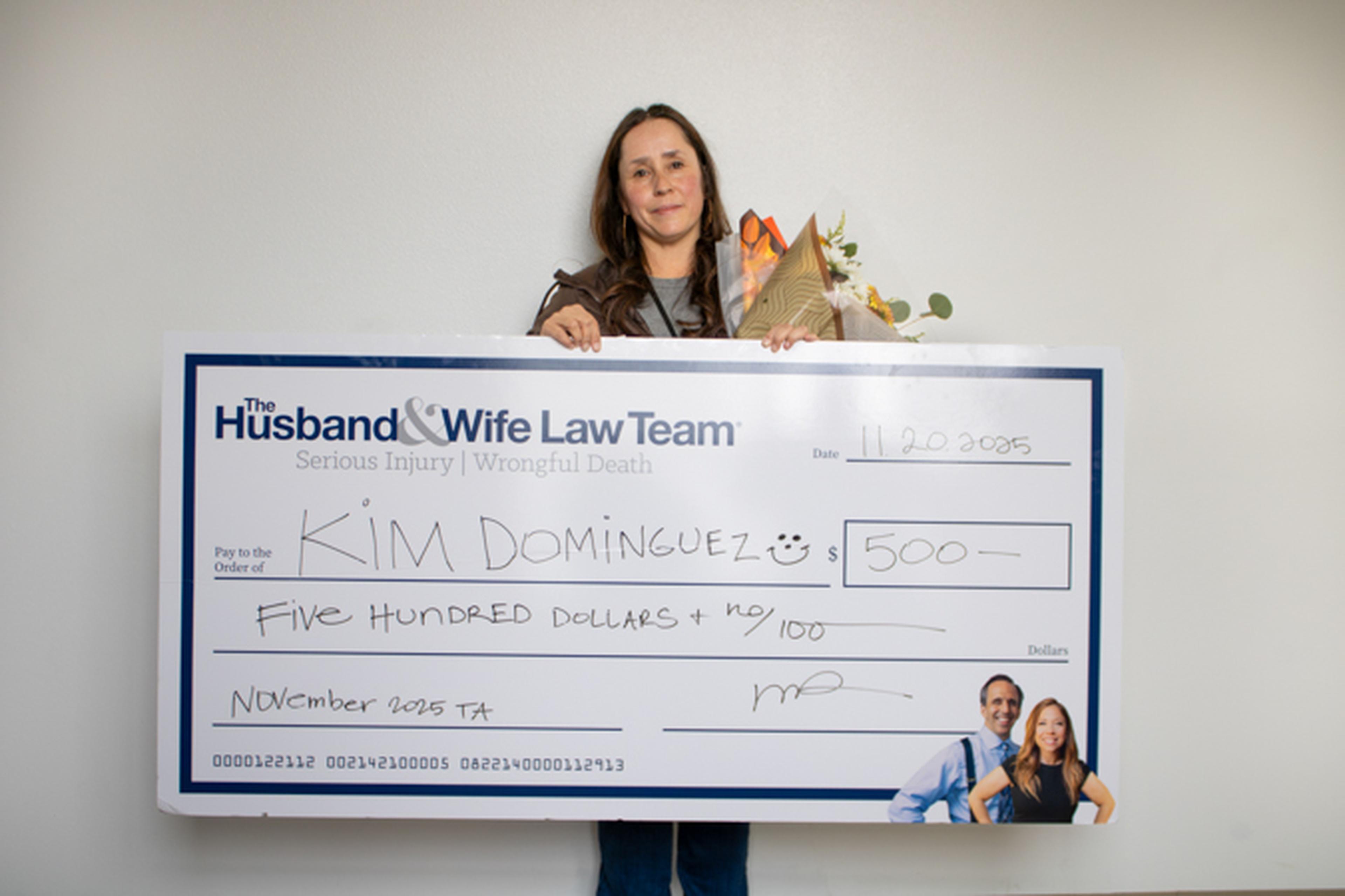 Woman holding bouquet and oversized check from The Husband & Wife Law Team made out to "Kim Dominguez" for $500.