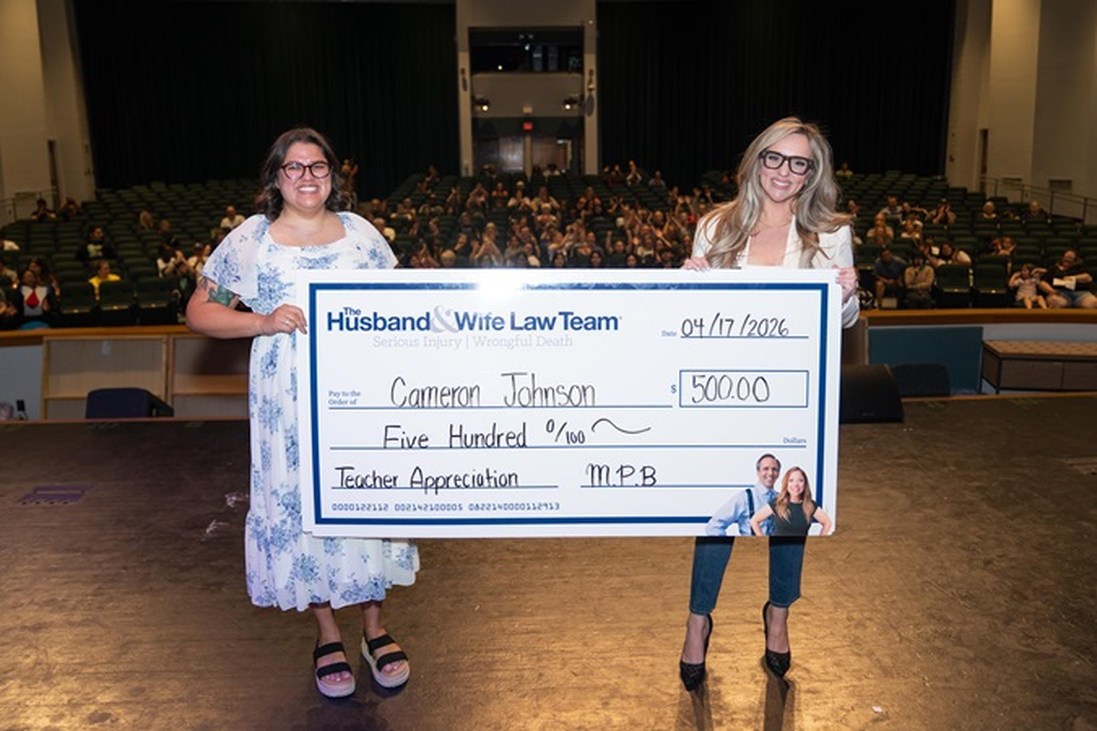 Jen and Ms. Johnson stand on stage holding a large $500 check, with the audience seated behind them.