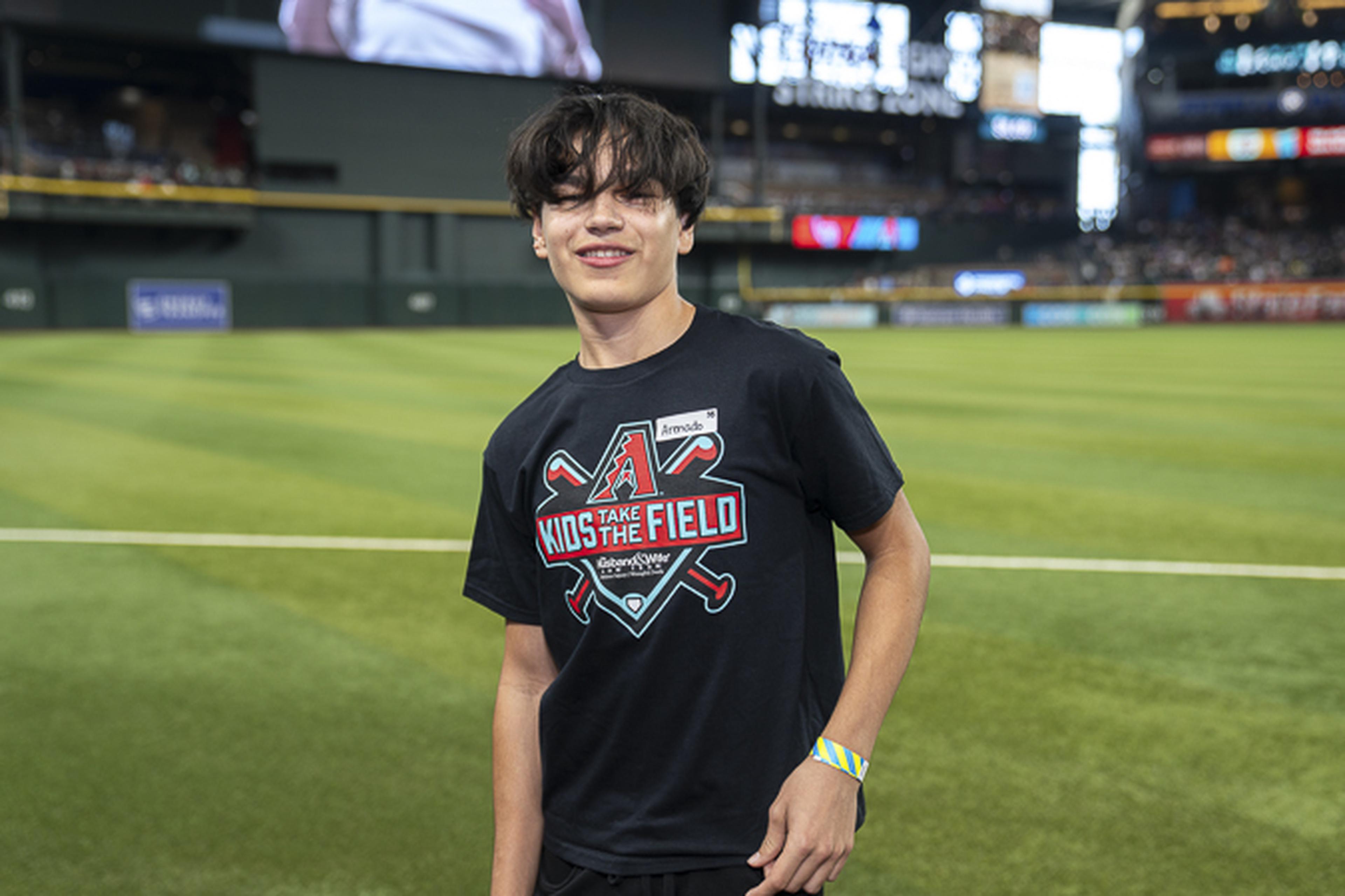 Smiling under the stadium lights, a young fan wearing a black “Take Kids to the Field” tee and wristband stands on the field with glowing screens behind them.