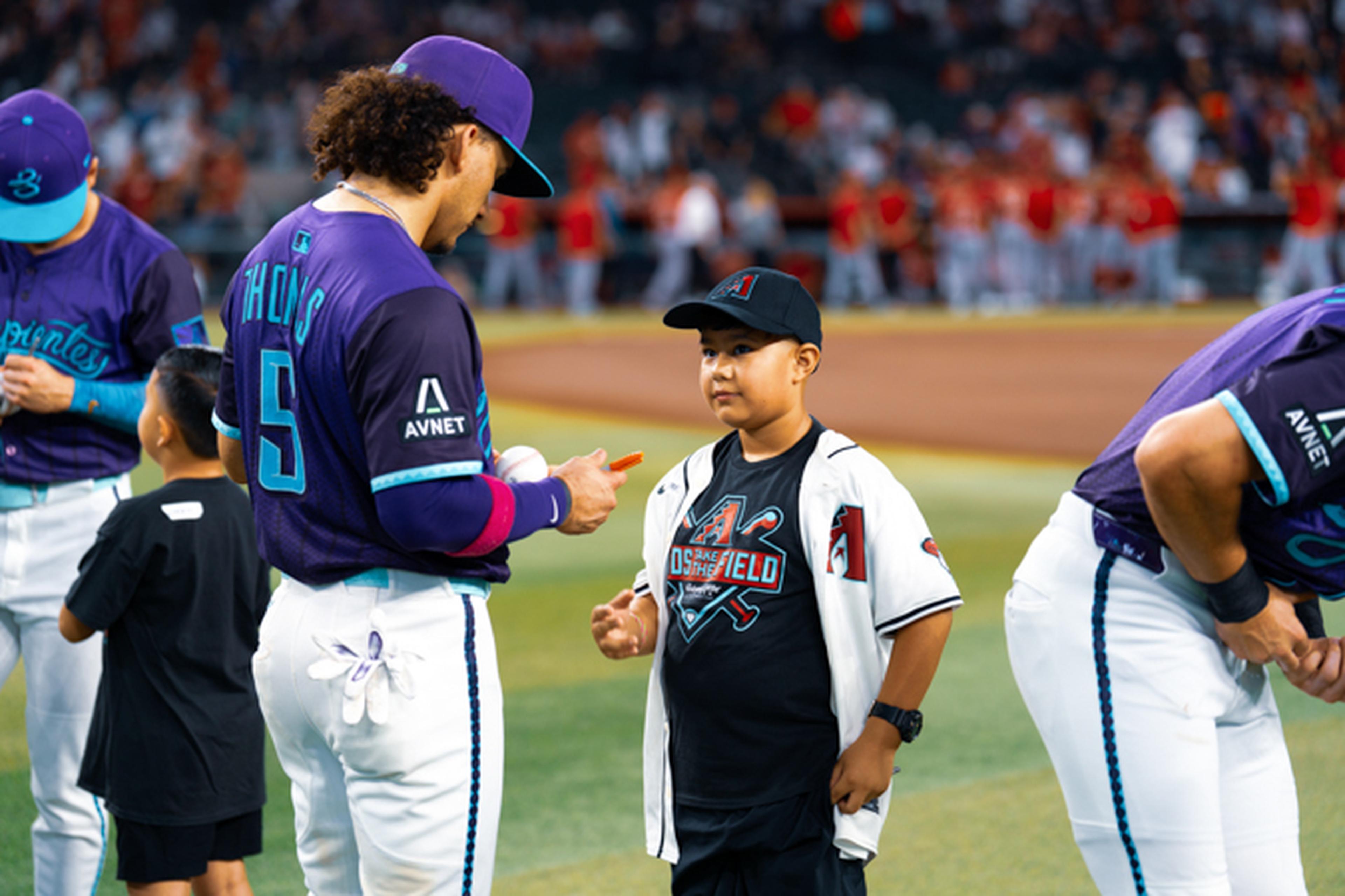 Baseball player in purple uniform signs an autograph for a young fan wearing a black and white jersey on a baseball field.