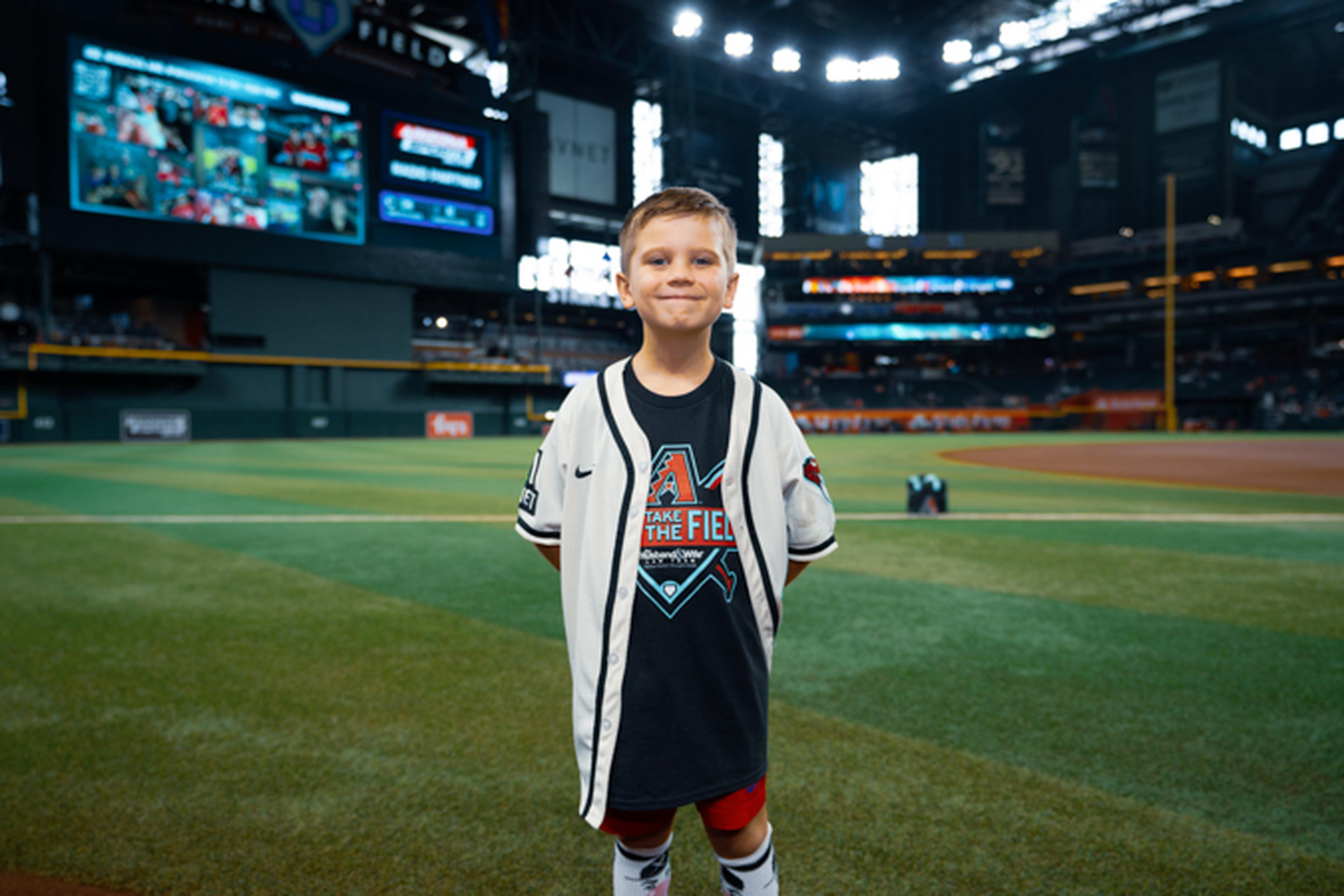Young boy in a baseball jersey stands on a professional baseball field, smiling. Stadium lights and scoreboard visible in the background.