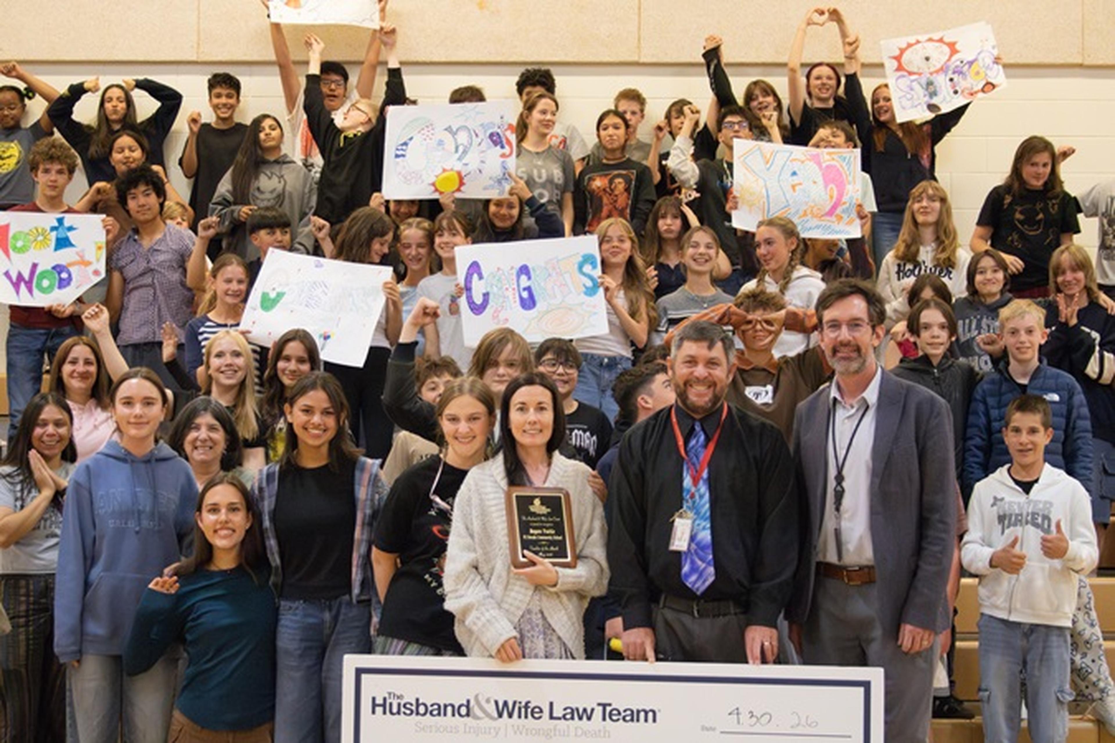 El Dorado Community School students and adults pose in the gym with colorful signs, a plaque, and a donation check from The Husband & Wife Law Team.