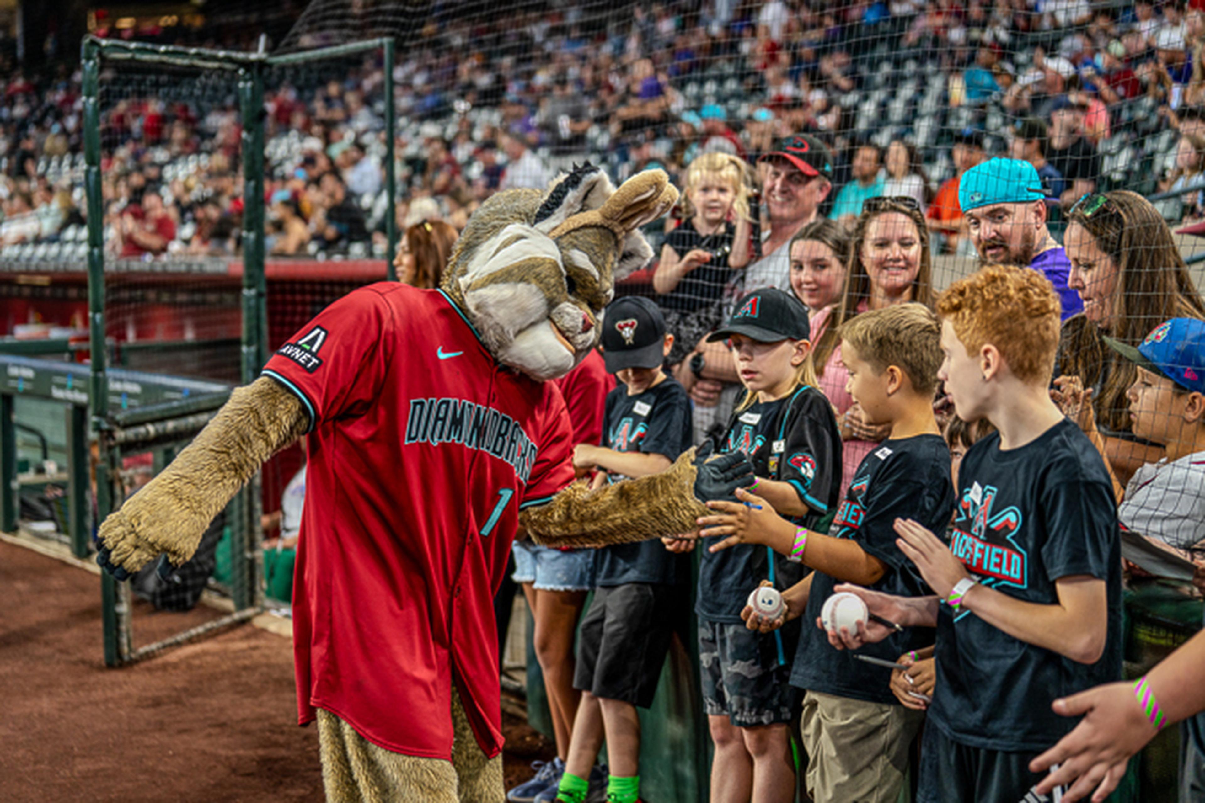 A sports mascot in a red jersey interacts with excited children and adults near the baseball field stands.