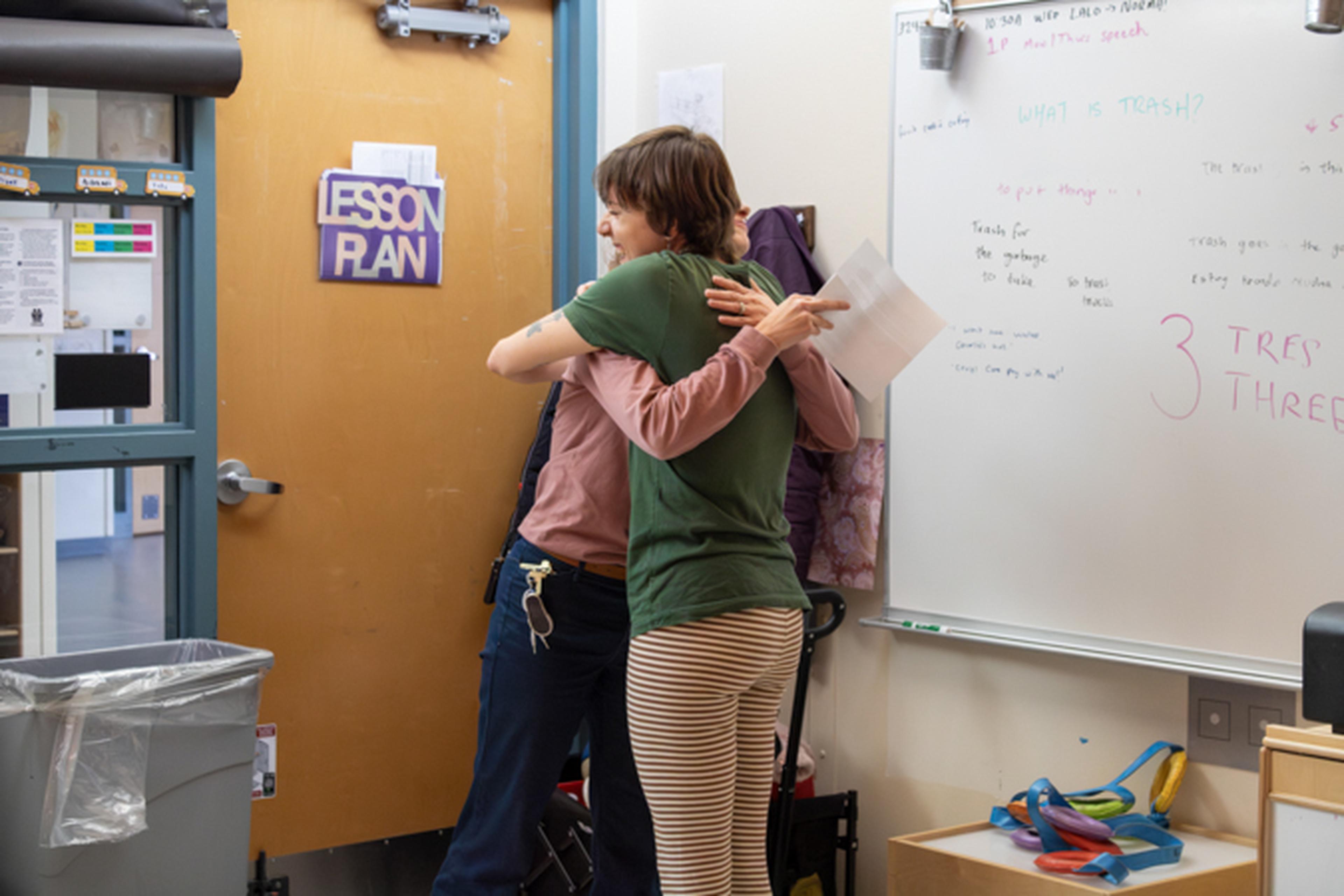 Two educators hug in a classroom doorway under a "Lesson Plan" sign, one holding papers.