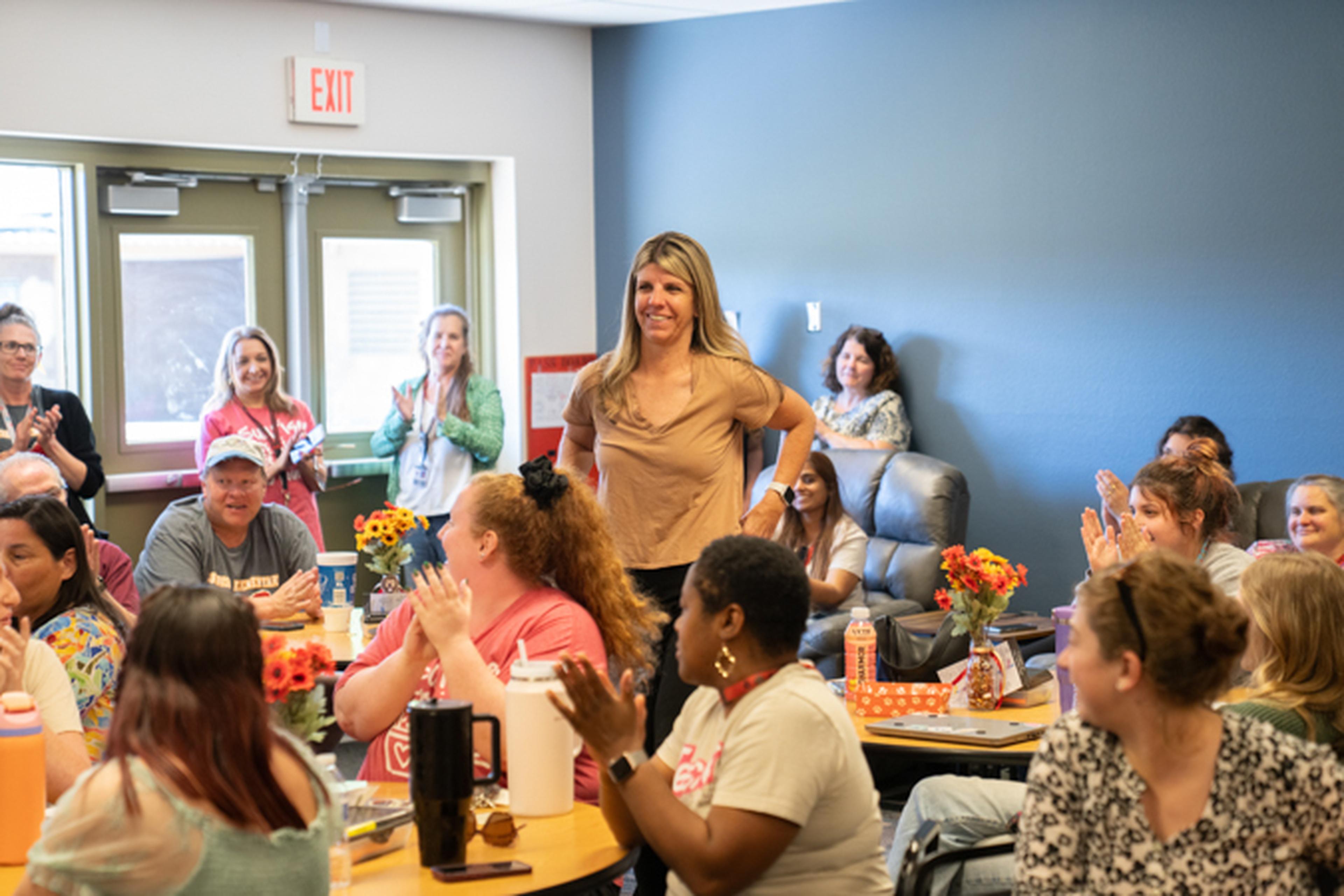 Ms. Shade stands smiling in a classroom, surrounded by applauding colleagues seated at tables with flowers and snacks.