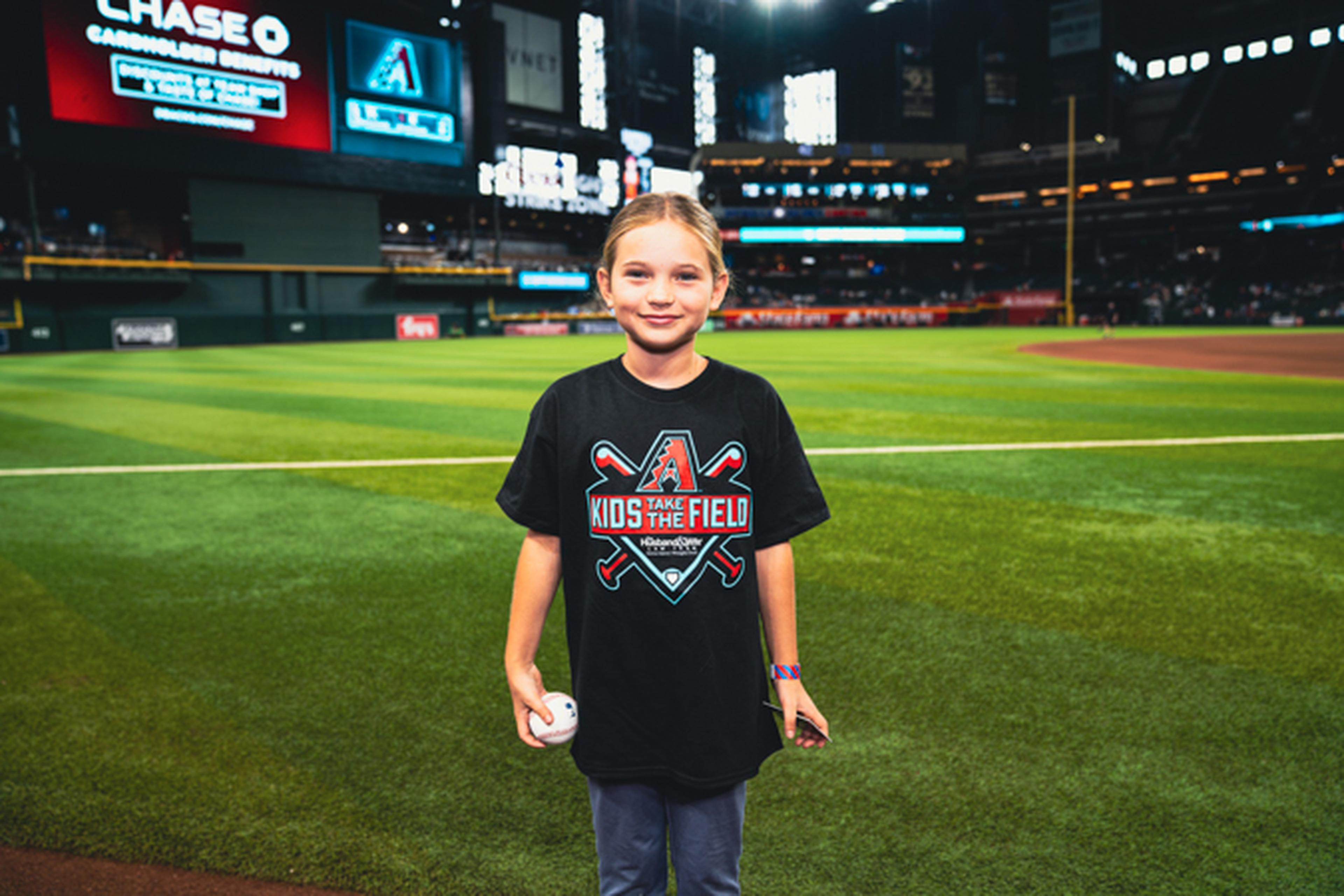 Young girl on a baseball field wearing a "Kids the Field" shirt, holding a baseball, with stadium lights and scoreboard in the background.