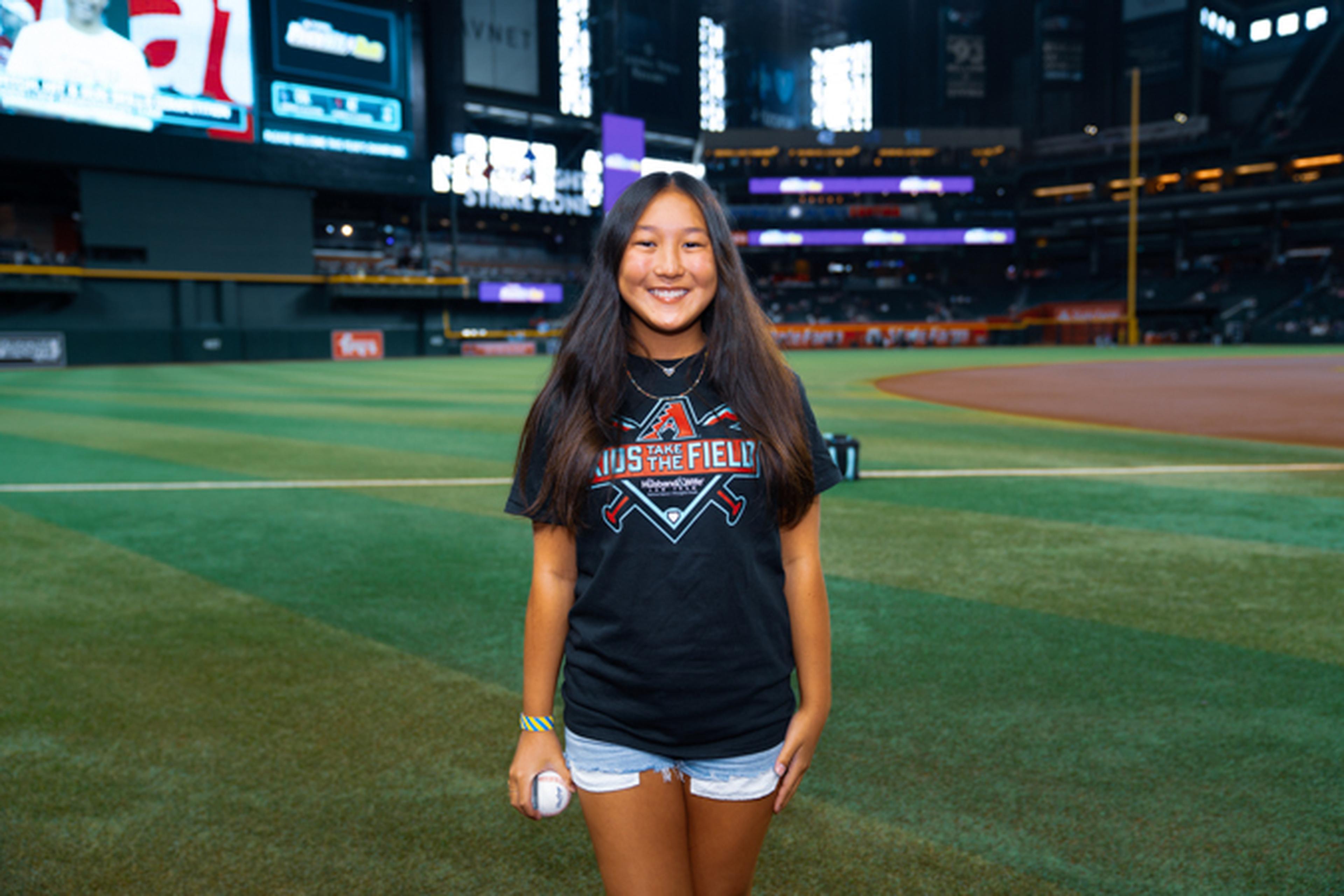 Smiling person with long hair stands on a baseball field holding a ball, wearing a black T-shirt and shorts. Stadium seats and screens in the background.