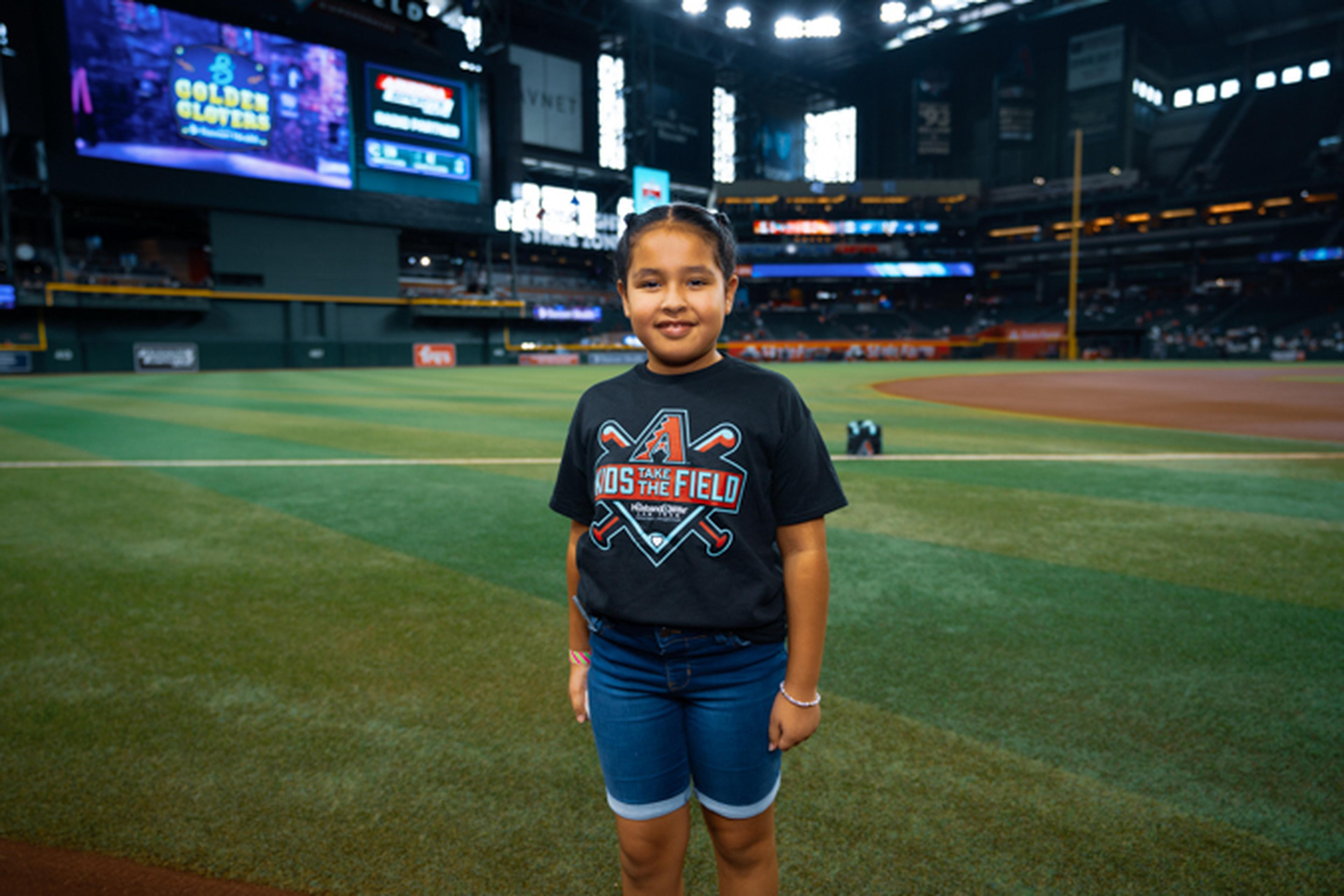 Young girl smiling on a baseball field, wearing a black t-shirt and blue shorts, with stadium lights and screens in the background.
