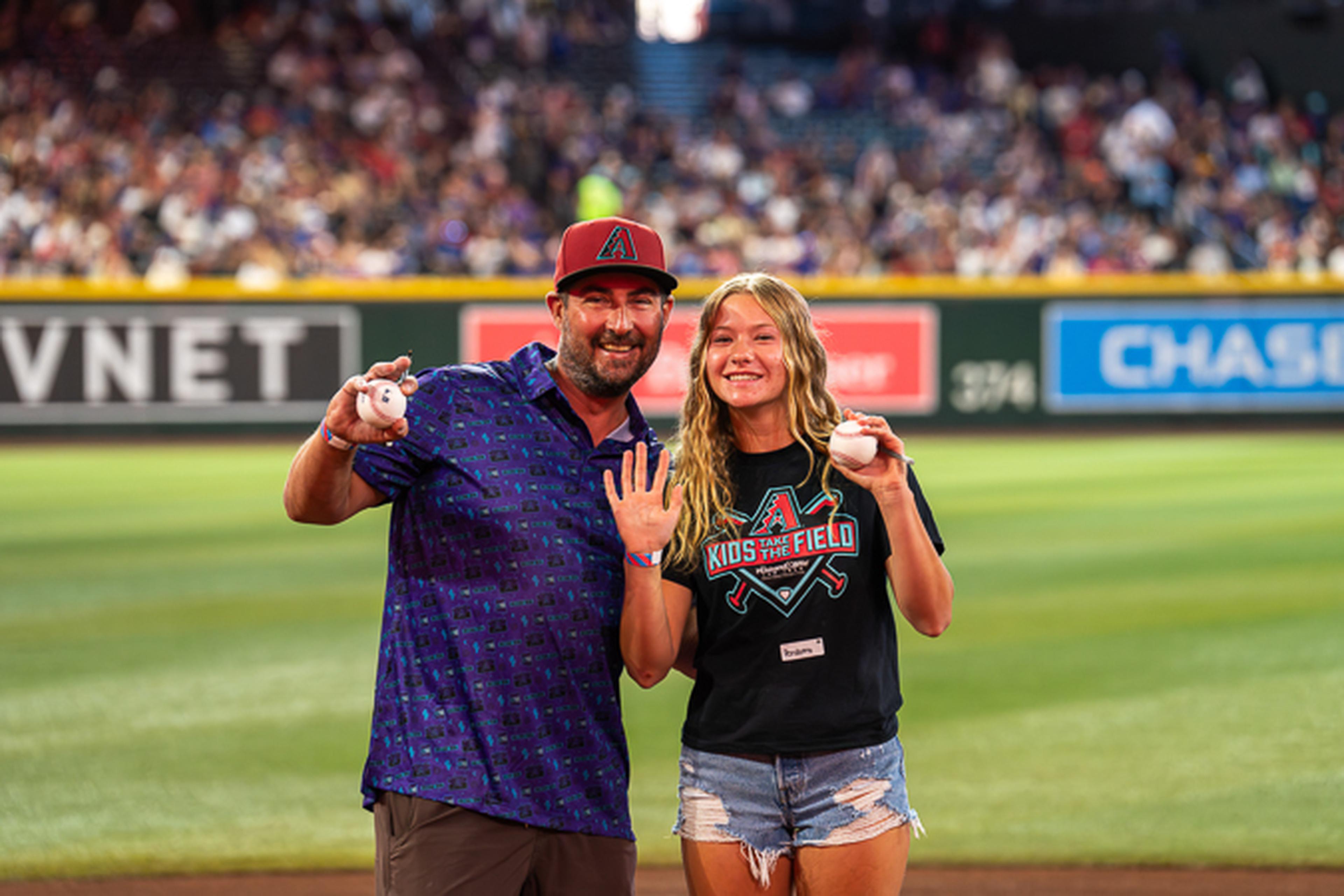 Father and daughter smile on Chase Field while holding baseballs, with her wearing a “Kids on the Field” shirt.