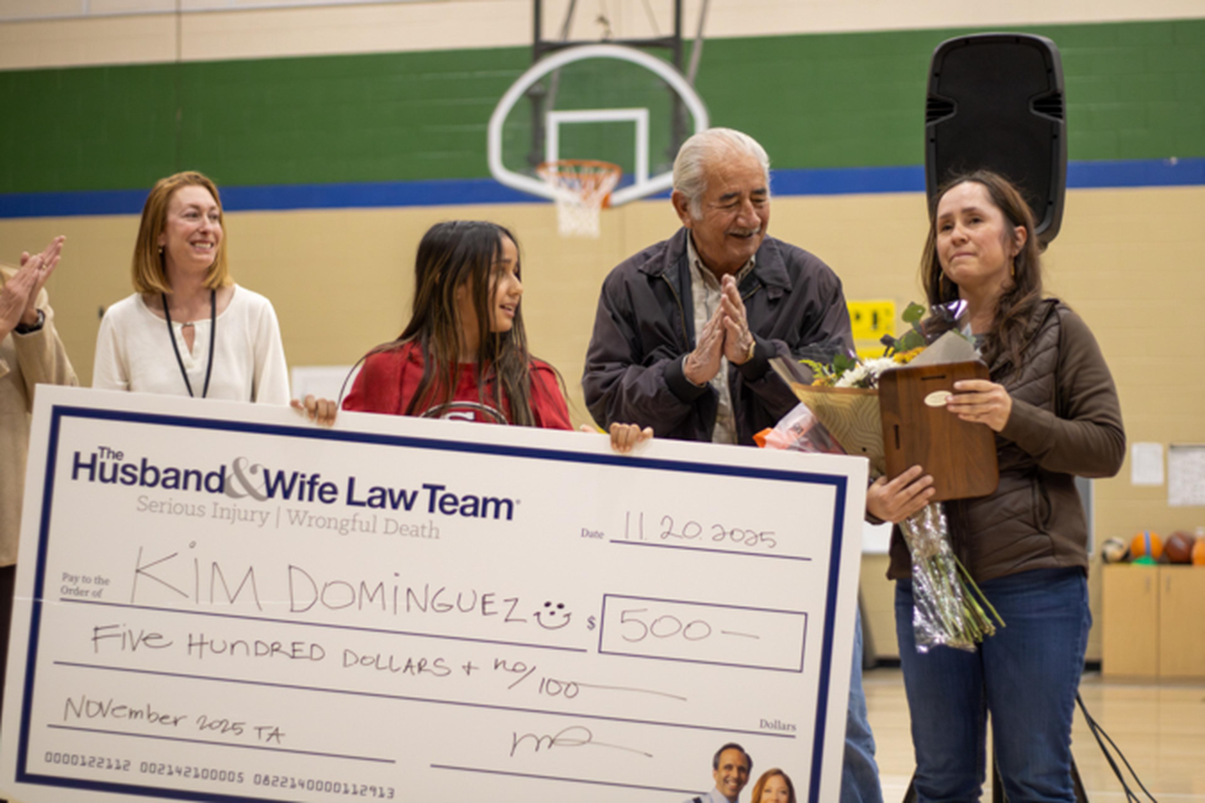 Young girl and adults in a gym holding an oversized $500 check as a woman holds flowers and others applaud.