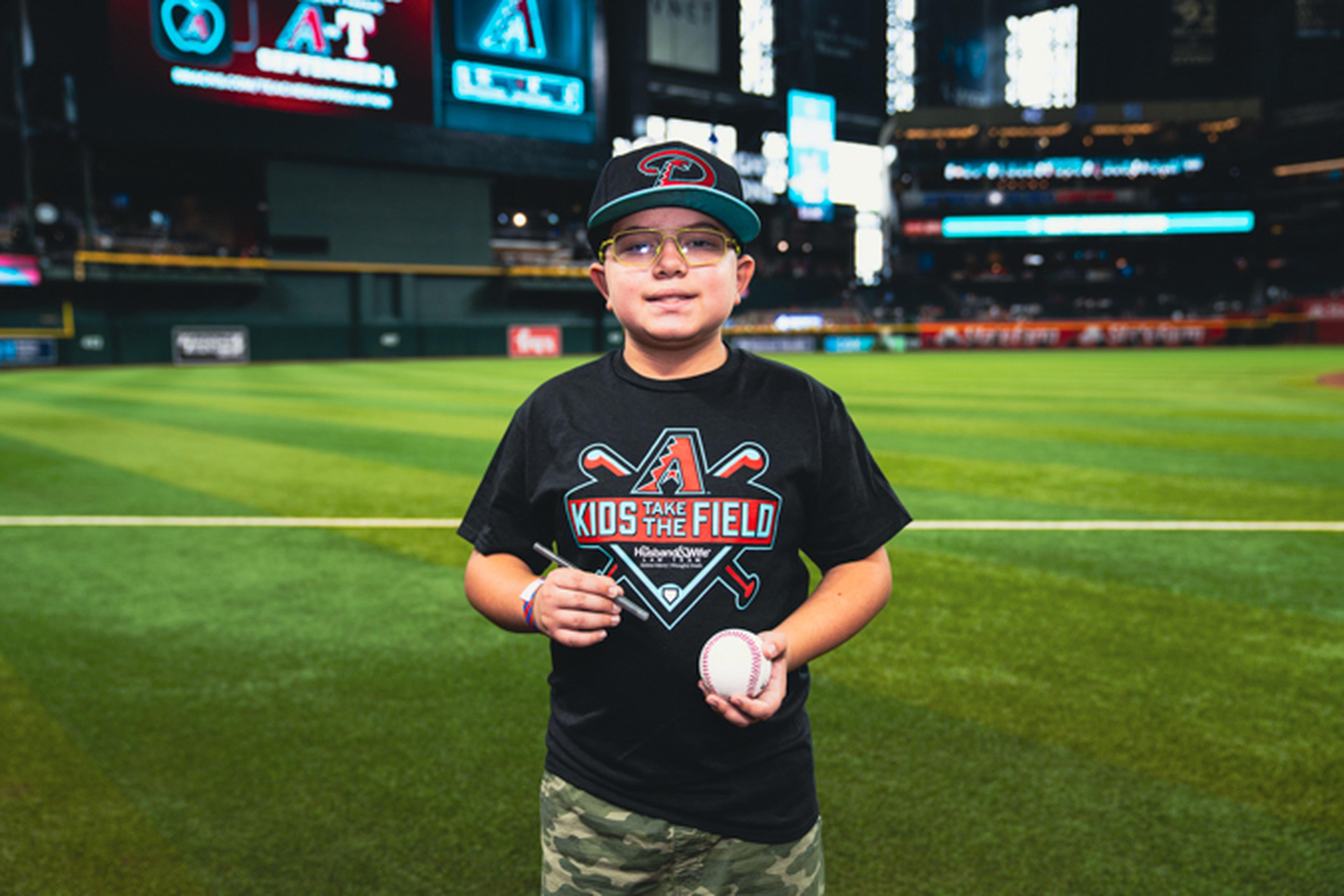A young person wearing a baseball cap and "Take Me to the Field" shirt holds a baseball on a stadium field with blurred stands in the background.