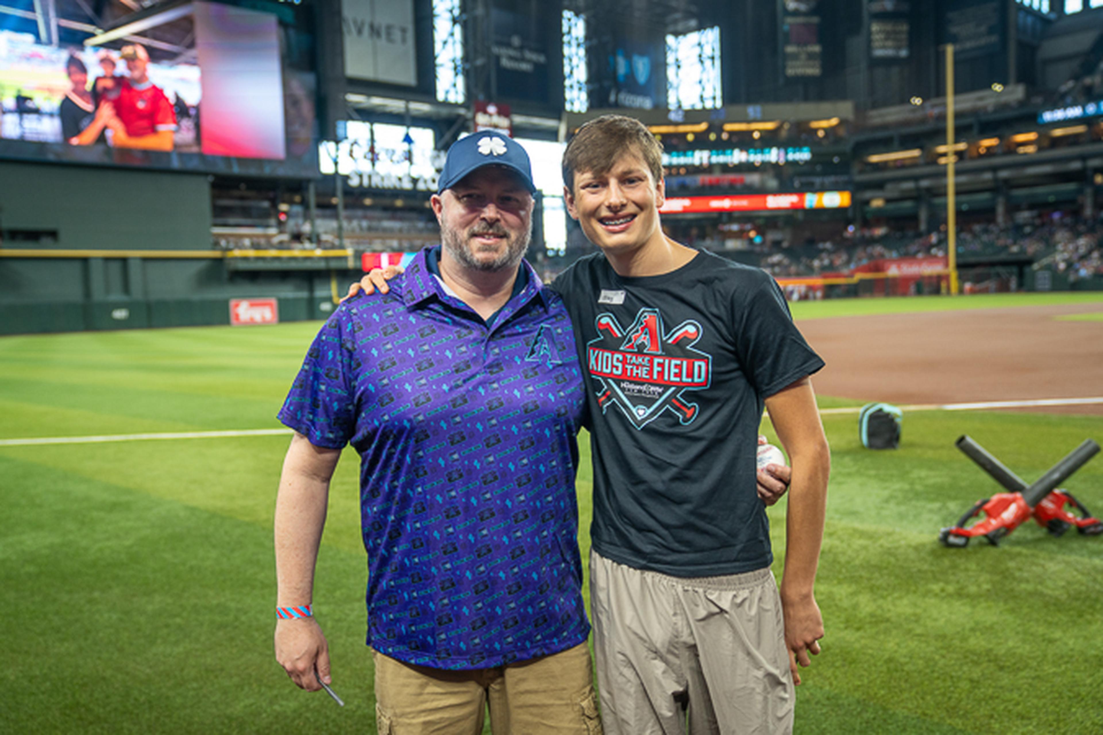 Father and son pose on the baseball field, one in a purple shirt and cap and the other in a black “Kids on the Field” shirt, with stadium seats and bat equipment behind them.