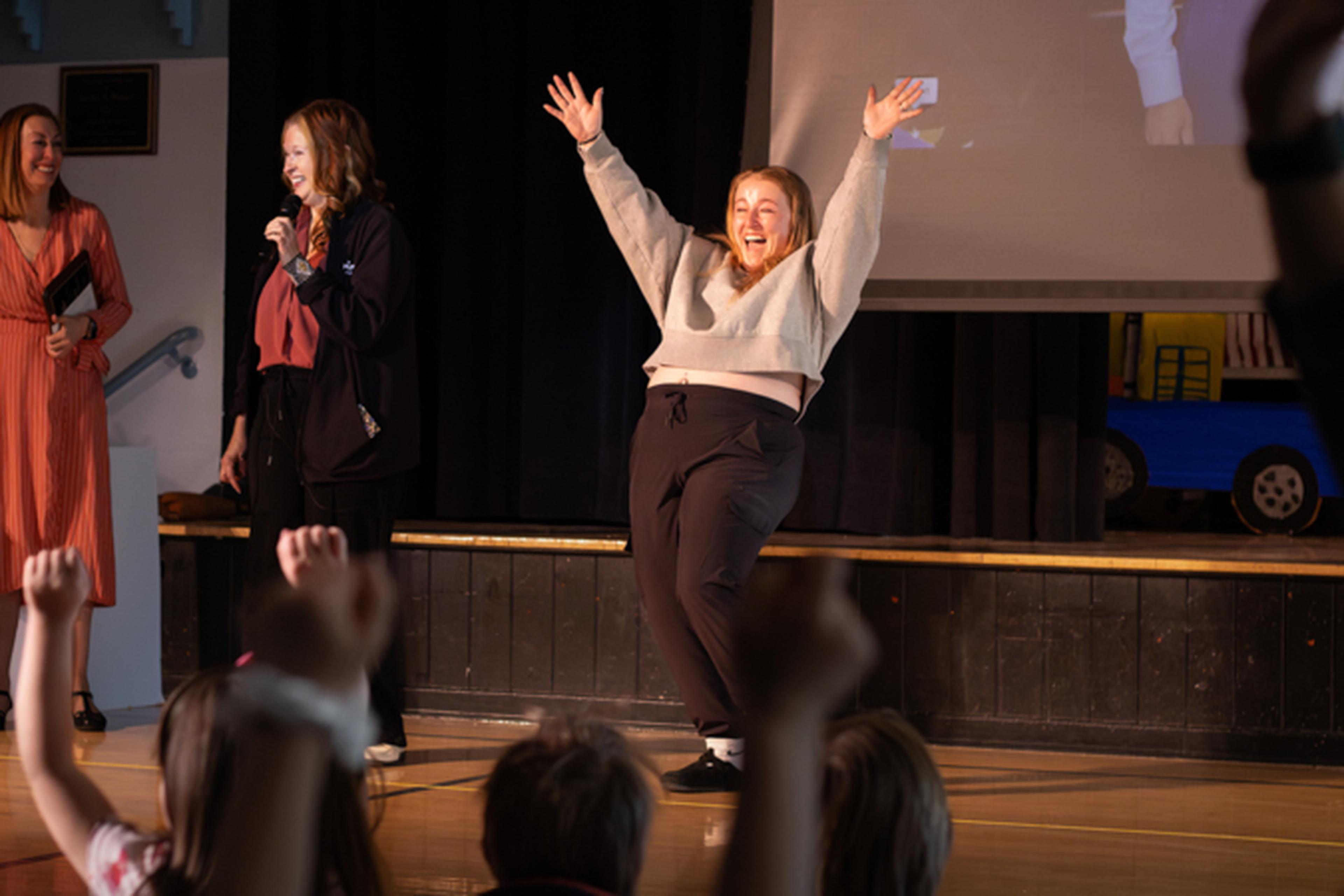 On stage at a school assembly, Ms. Amy lifts her arms triumphantly while Lisa from The Husband & Wife Law Team cheers her on for her accomplishments.